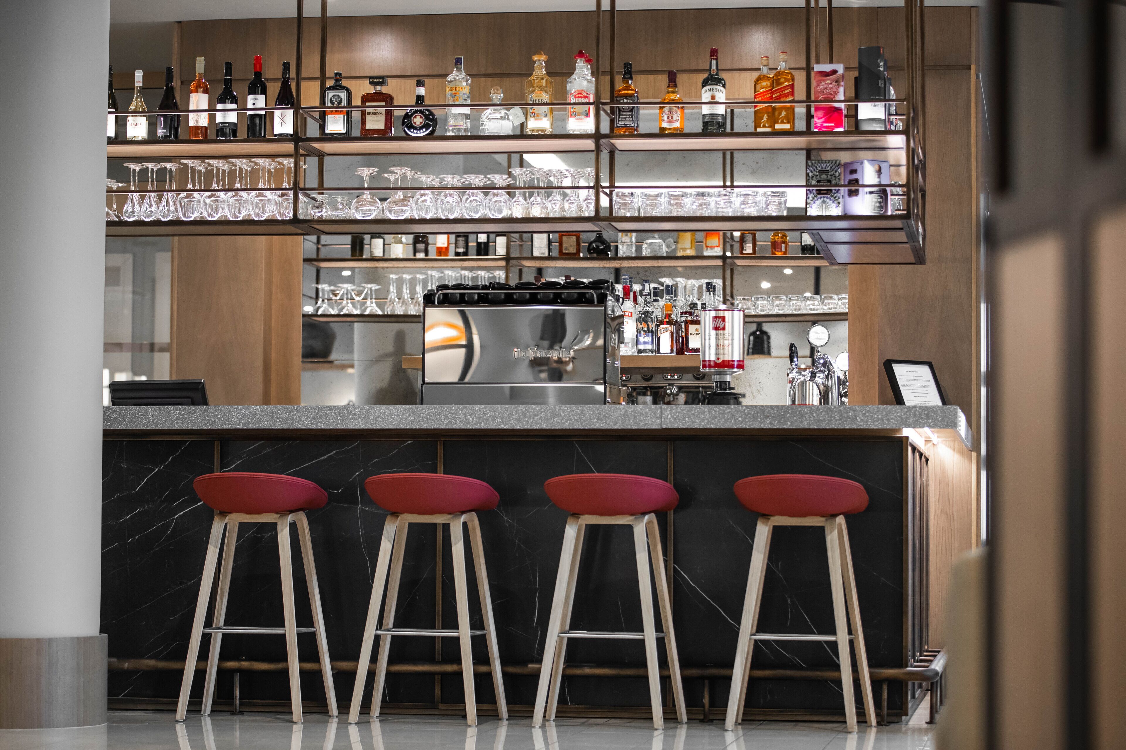 Una zona de bar con mostrador de mármol, tres taburetes rojos y estanterías llenas de botellas y vasos.