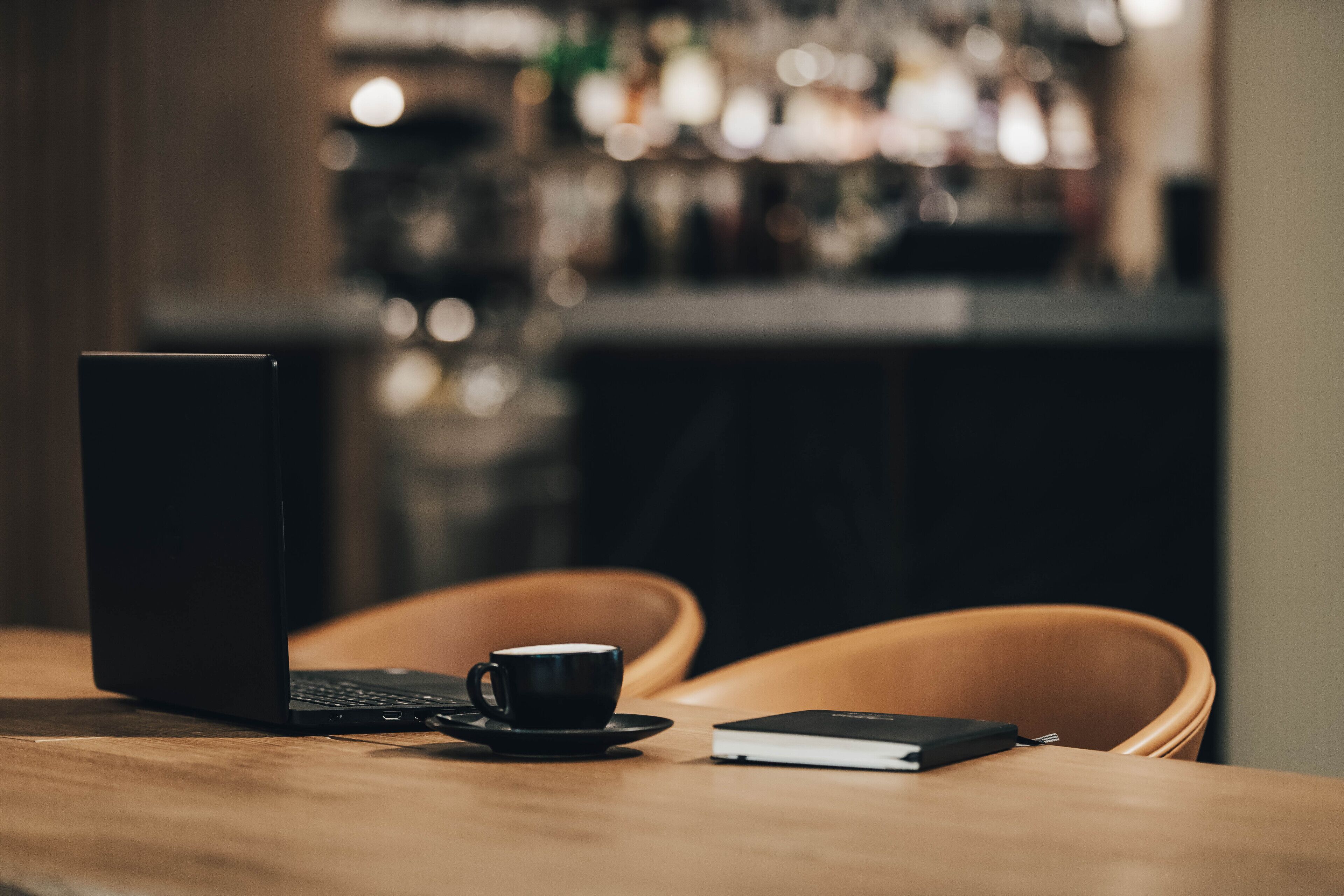 A laptop, a cup of coffee, and a notebook on a wooden table in a bar setting.