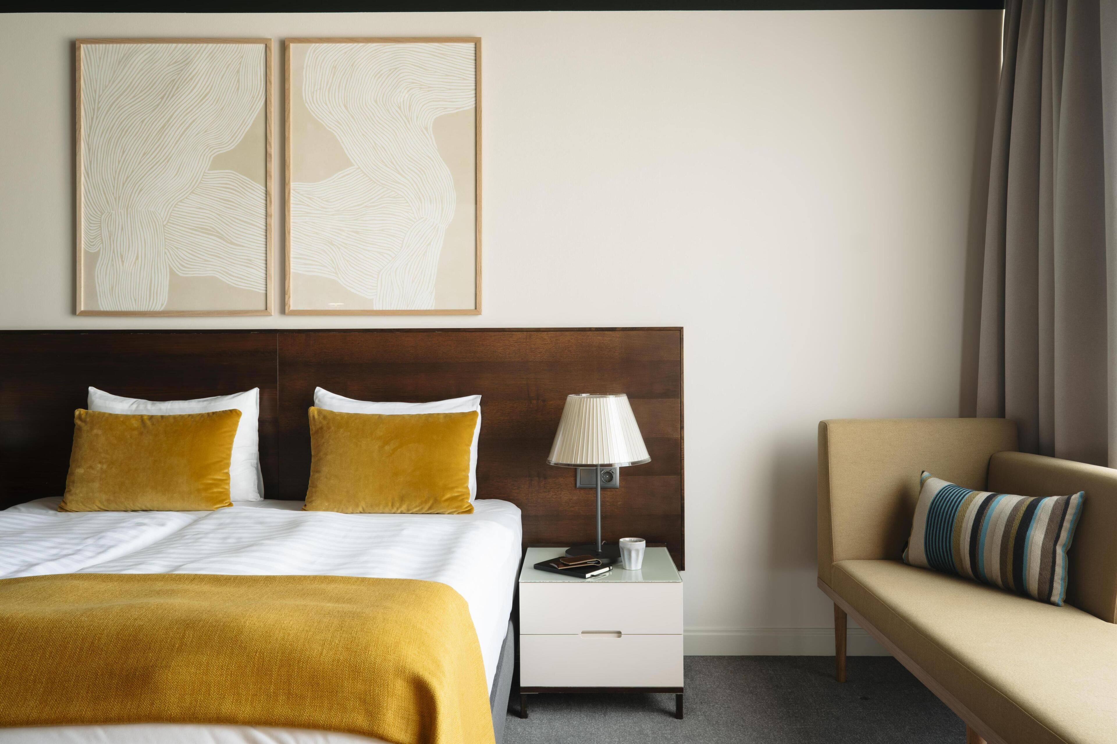 A hotel room with a bed, a nightstand, a lamp, a sofa, and two abstract paintings on the wall.