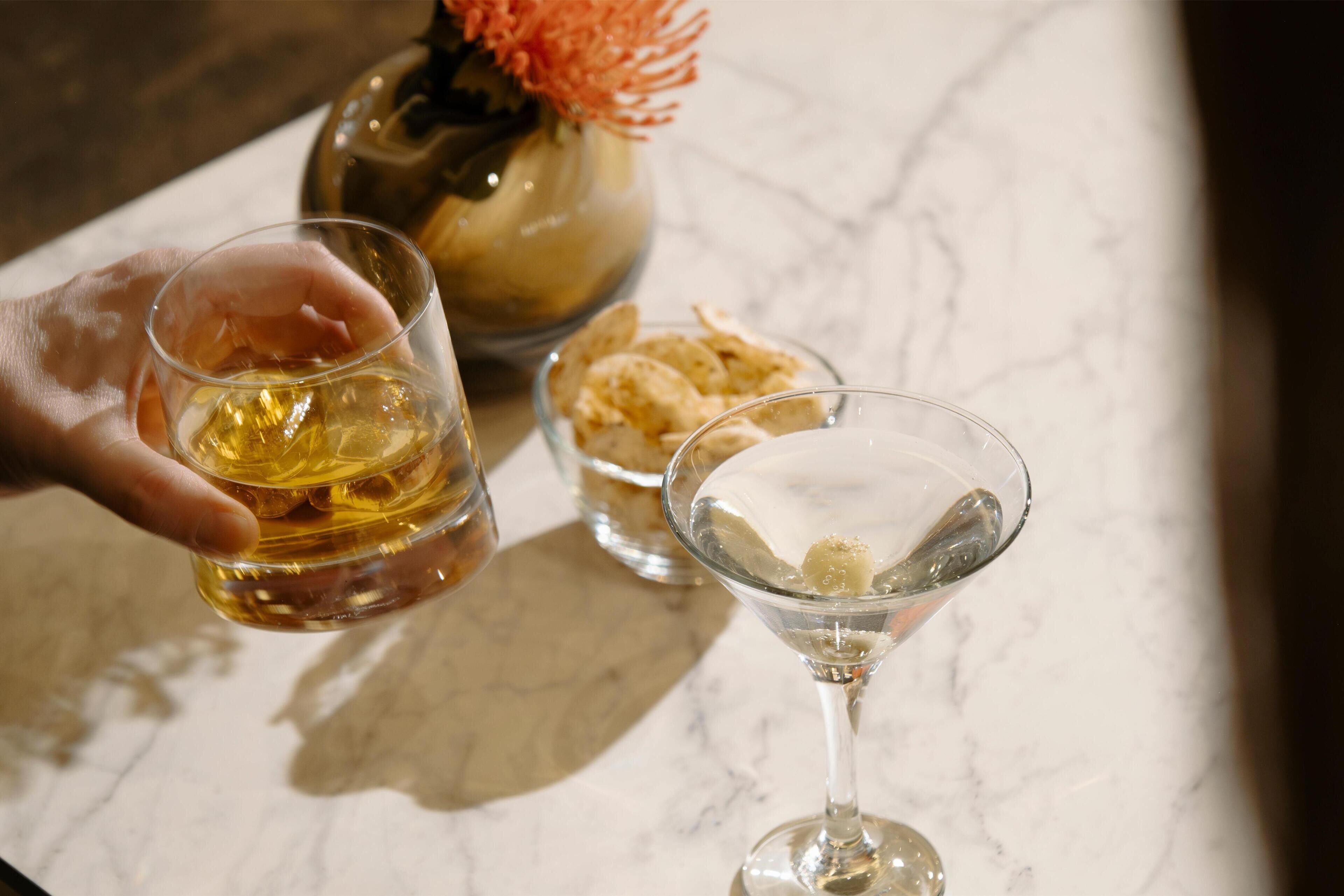 Una mano sostiene un vaso con líquido ámbar, un vaso de martini con líquido transparente y hielo, y un cuenco pequeño con aperitivos sobre una superficie de mármol.