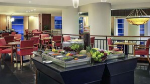 Comedor de hotel con sillas rojas, un bufé de frutas y verduras, e iluminación moderna.