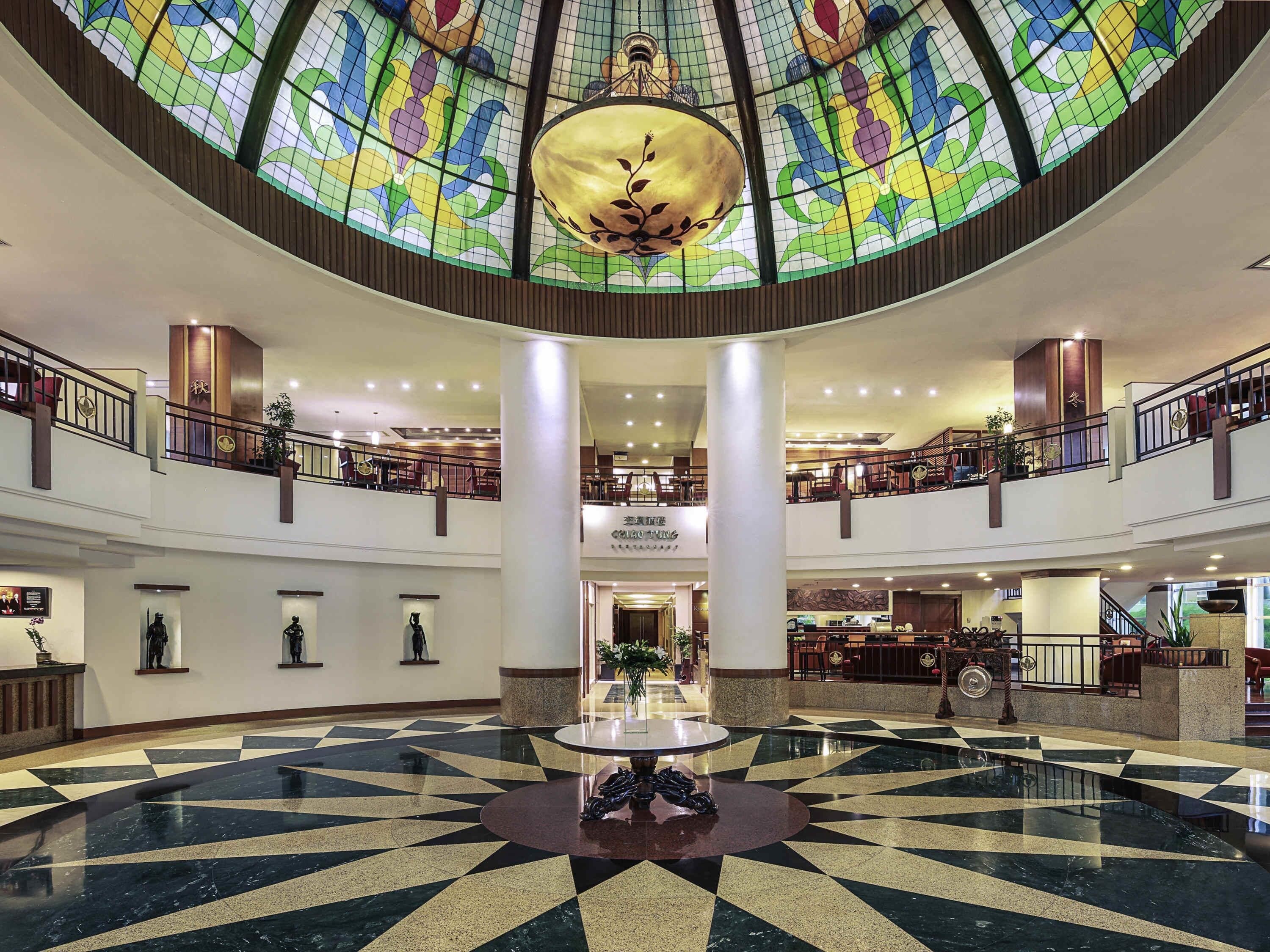 A hotel lobby with a central circular seating area, a high ceiling with a stained glass dome, multiple floors with railings, and a visible restaurant.
