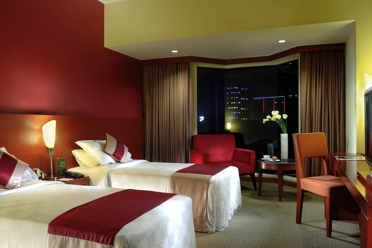 Une chambre d’hôtel avec deux lits, un fauteuil rouge, une petite table sur laquelle est posé un vase de fleurs, et une vue sur la ville la nuit.
