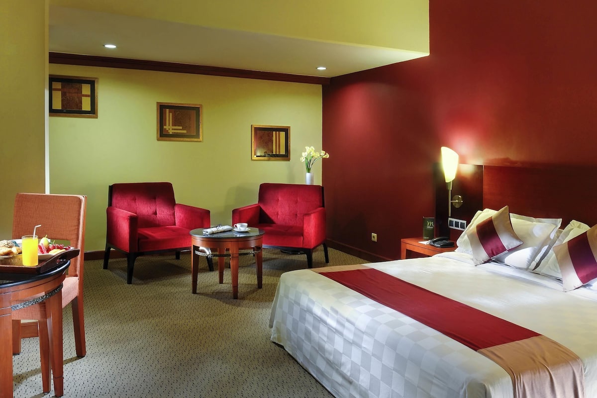 Une chambre d’hôtel avec un lit, deux fauteuils rouges, une petite table avec une tasse et un panier de fruits, et un vase avec des fleurs accroché au mur.