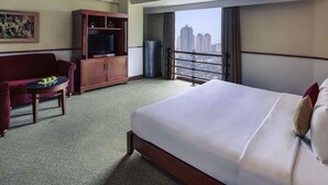 Habitación de hotel con una cama grande, televisión, zona de estar con sofá y una mesita, y balcón con vistas a la ciudad.