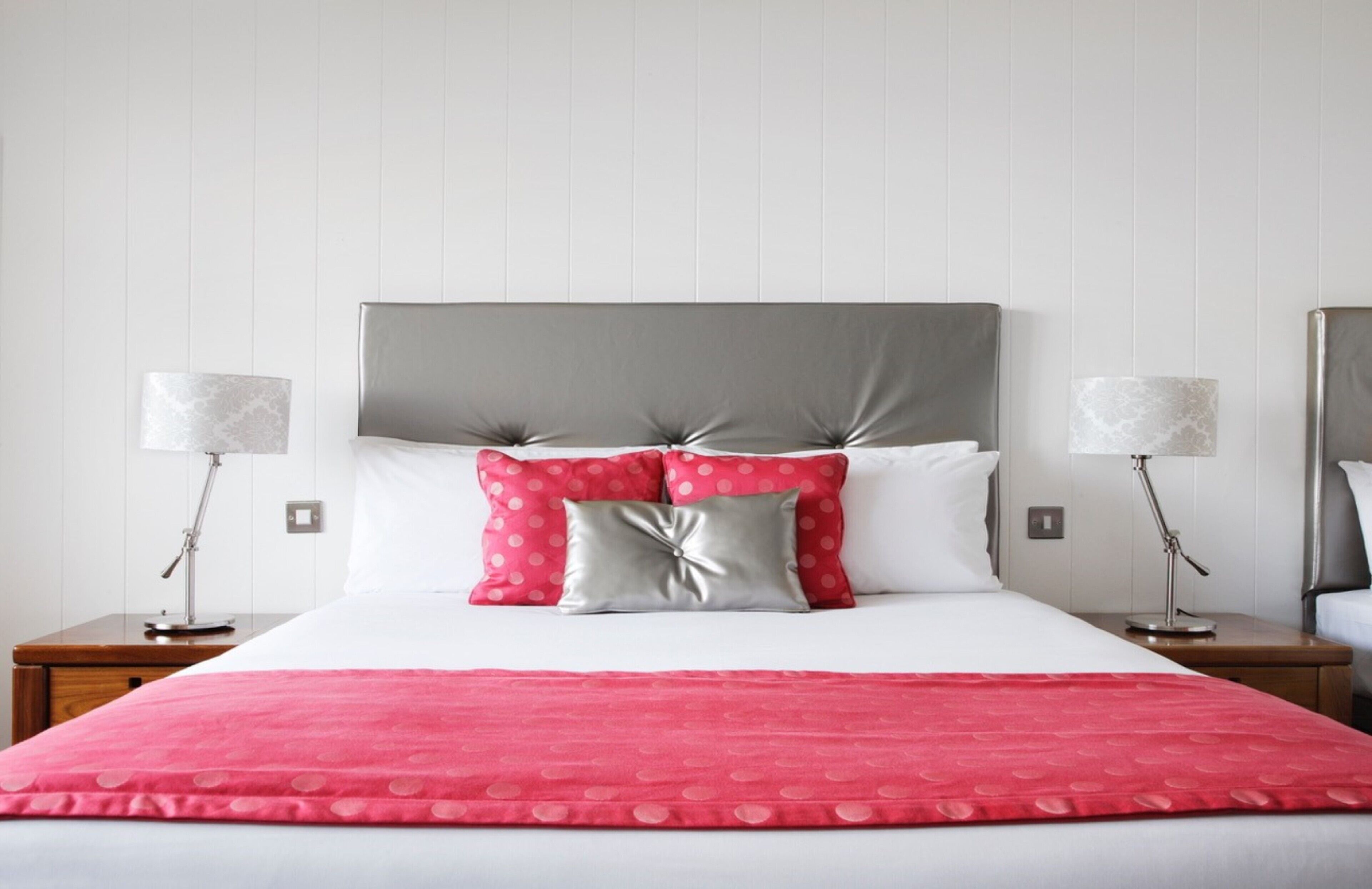 A neatly made bed with a grey headboard, white pillows, and a red polka dot bedspread. There are two bedside tables with lamps on either side of the bed.