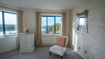 A room with a view of a lake, a white armchair, a cabinet with a vase of flowers, and a mirror on the wall.