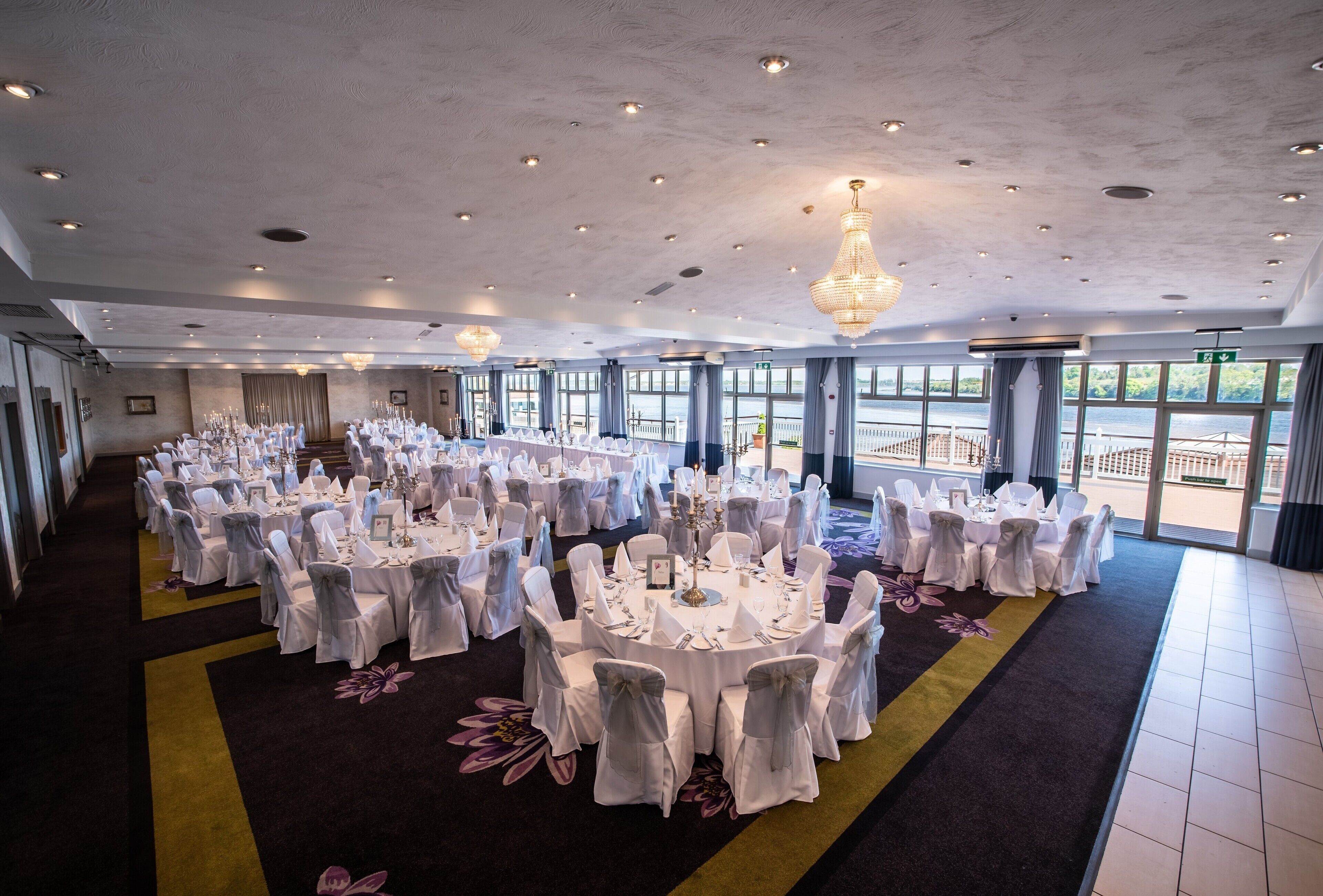 A large banquet hall with round tables set for a formal event, featuring chandeliers and large windows overlooking a scenic view.