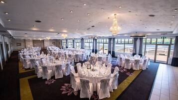 A large banquet hall with round tables set for a formal event, featuring chandeliers and large windows overlooking a scenic view.