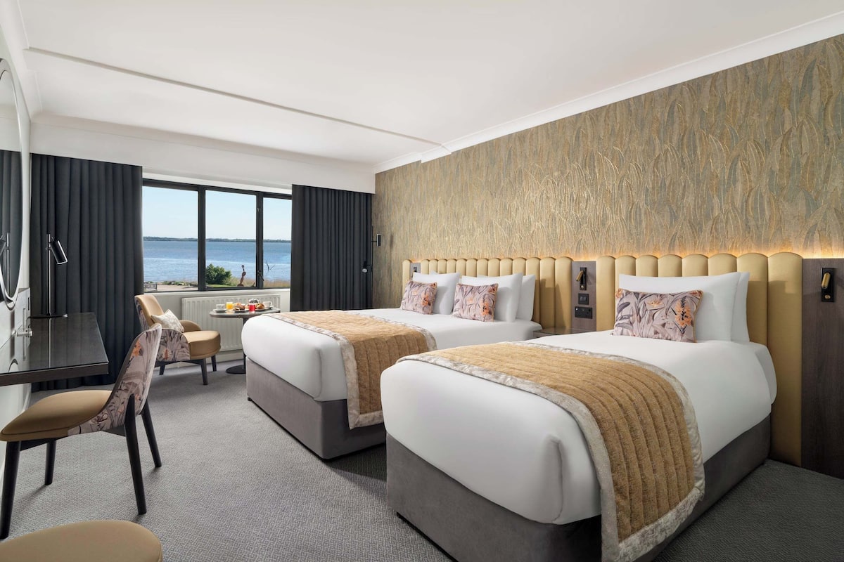 A hotel room with two beds, a desk, a chair, and a view of the sea.