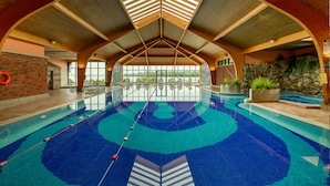 Una piscina interior con suelo de baldosas azules y blancas, vigas de madera en el techo y una pared de piedra en un lateral.