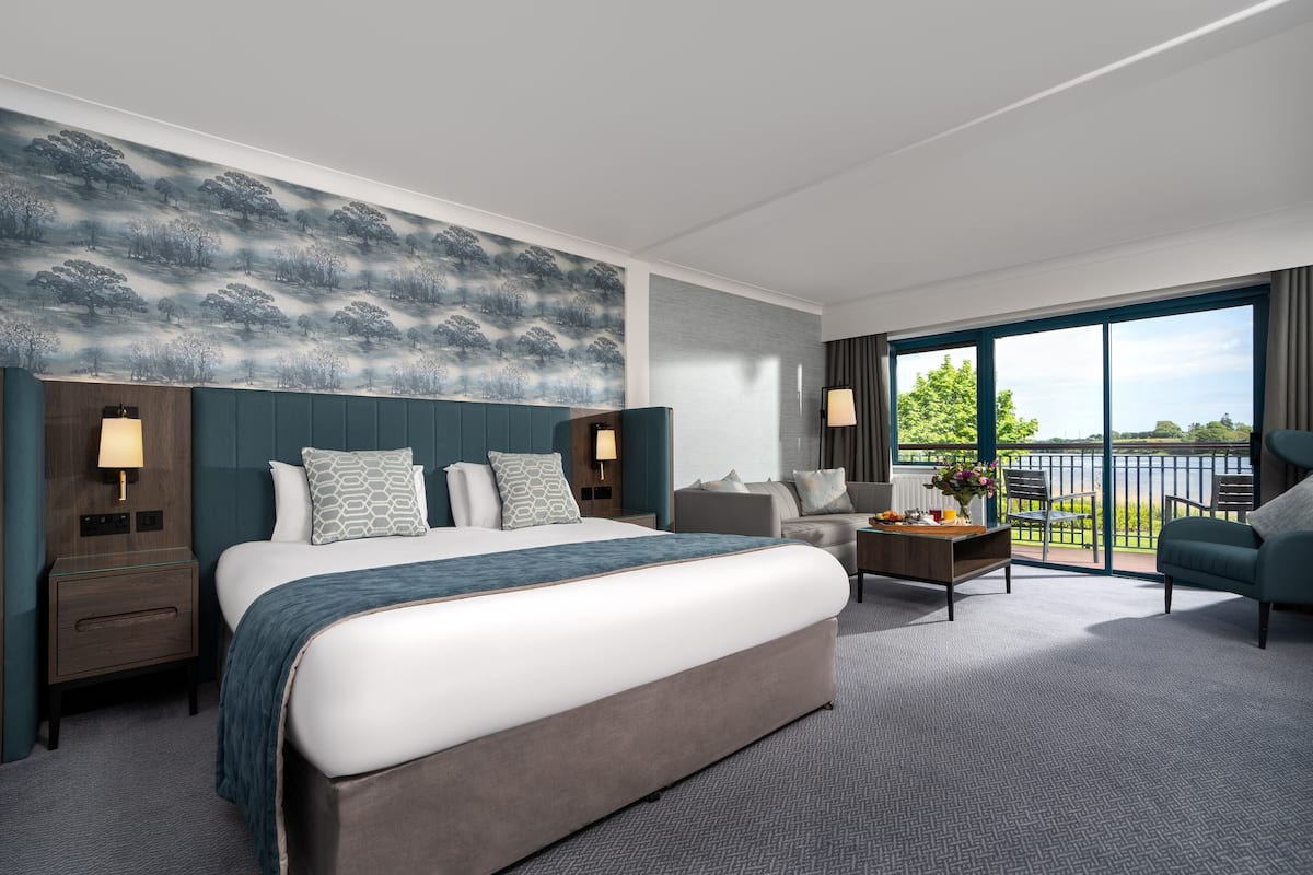 A modern hotel room with a large bed, a sofa, a small table, and a balcony with a view.