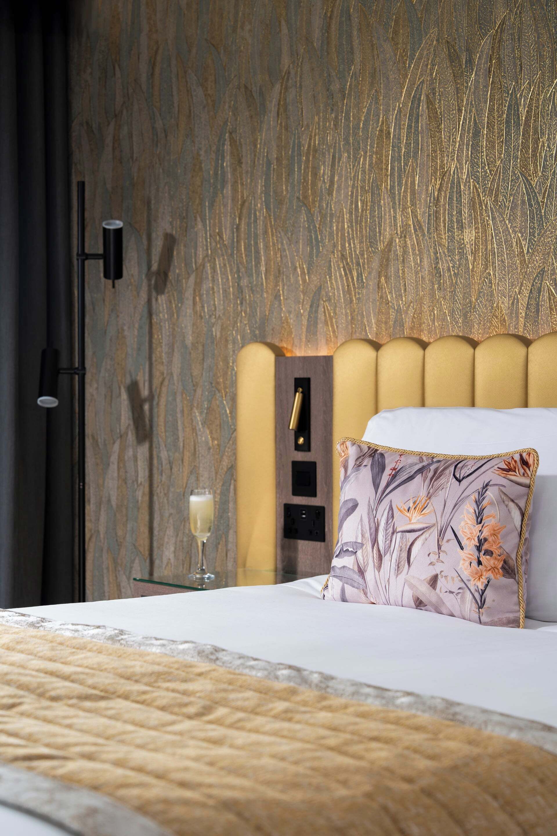 Cama arrumada com travesseiro floral, luminária de cabeceira e uma taça de vinho no criado-mudo.