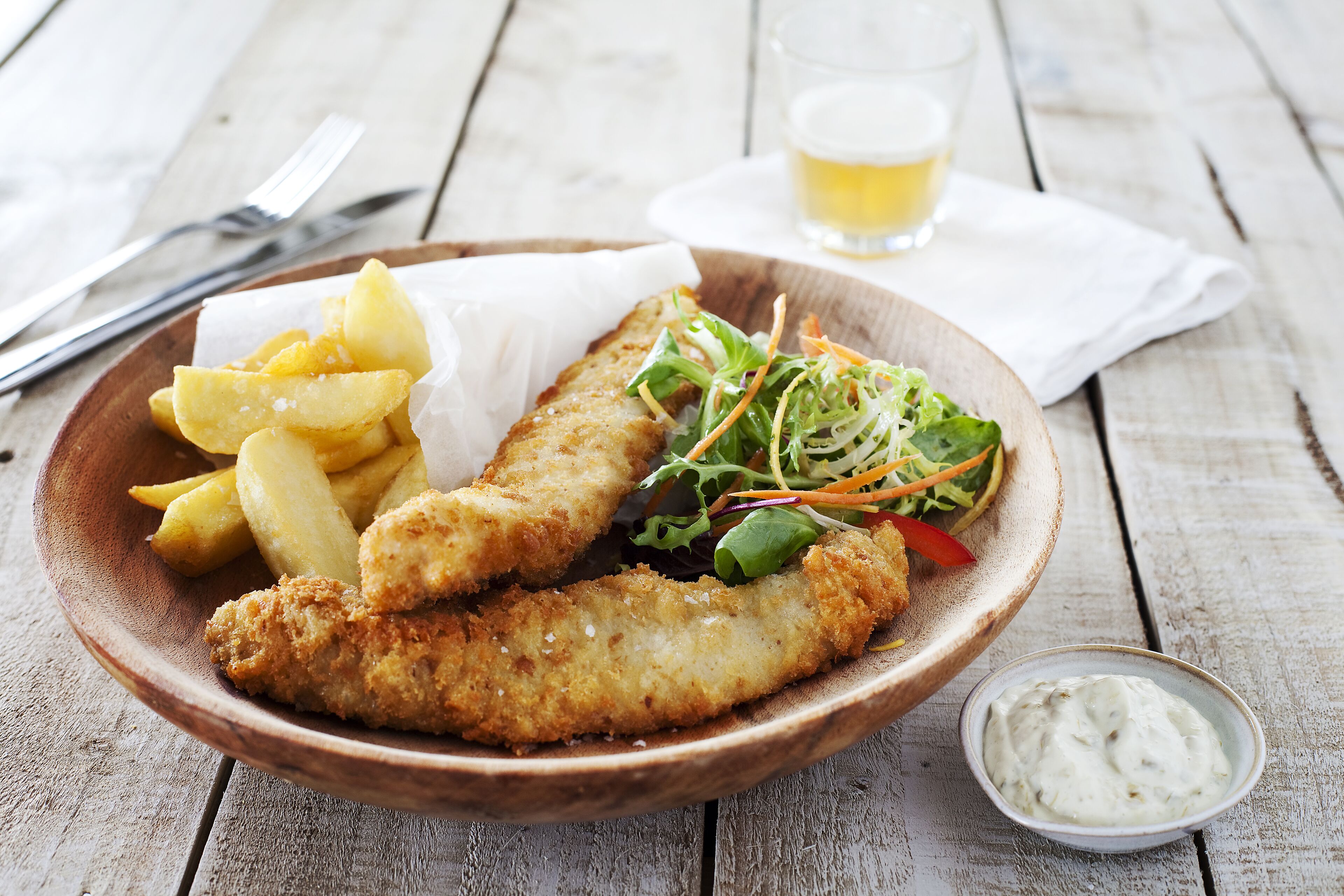 Un plato de filetes de pescado empanizados, papas fritas y una ensalada con un recipiente pequeño de salsa, servido sobre una mesa de madera junto a una cerveza y cubiertos.