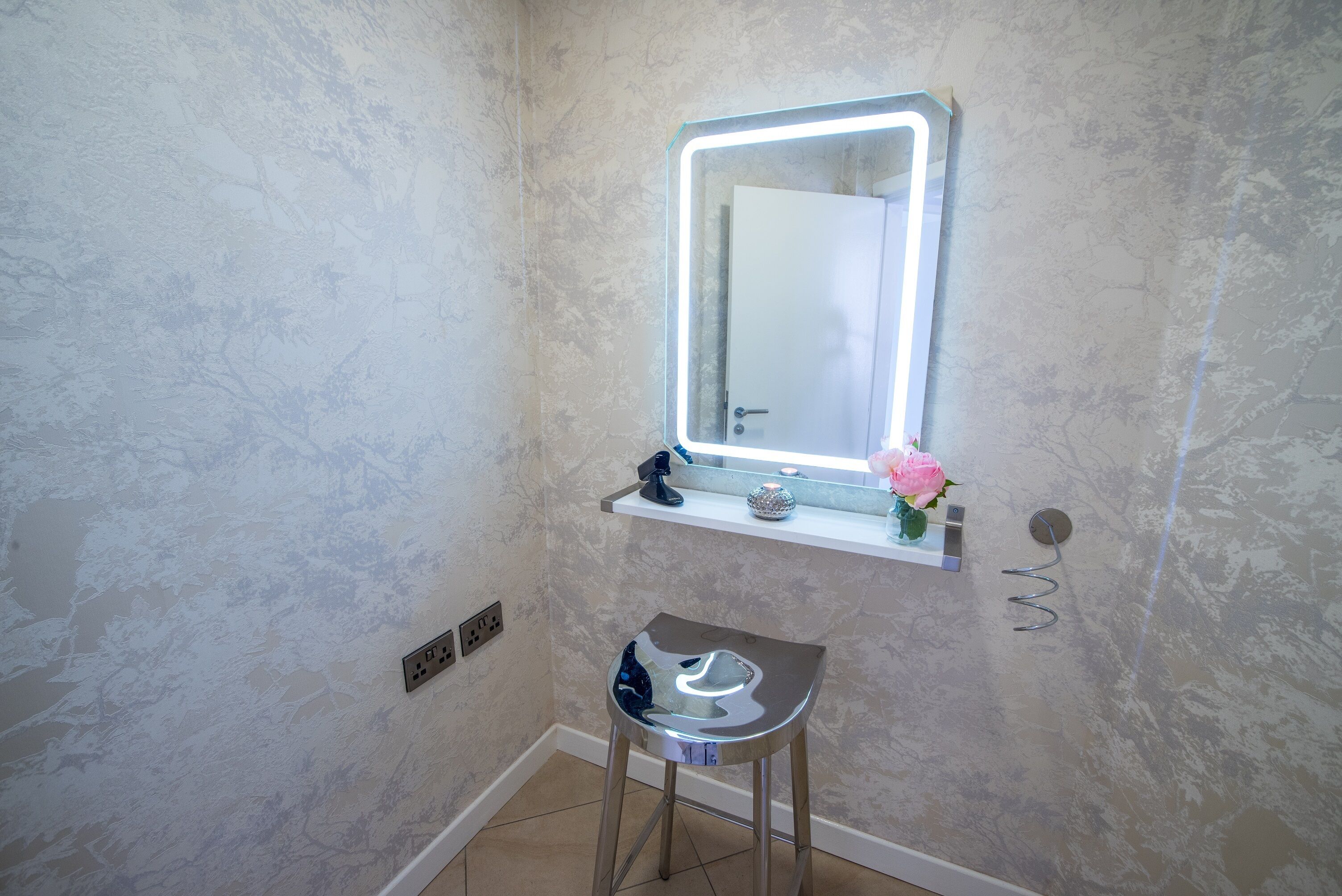 Un baño con pared de mármol, tocador con espejo, un taburete pequeño y una repisa con una flor.