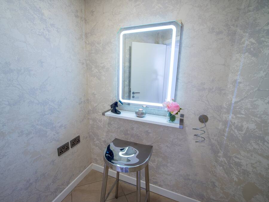 A bathroom with a marble wall, a vanity with a mirror, a small stool, and a shelf with a flower.