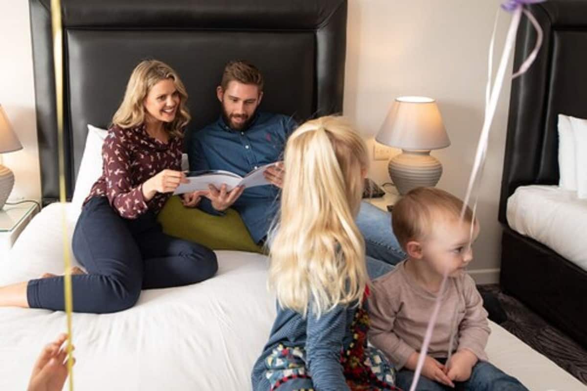 A family of four on a bed with a black headboard, a bedside lamp, and a nightstand with a lamp.