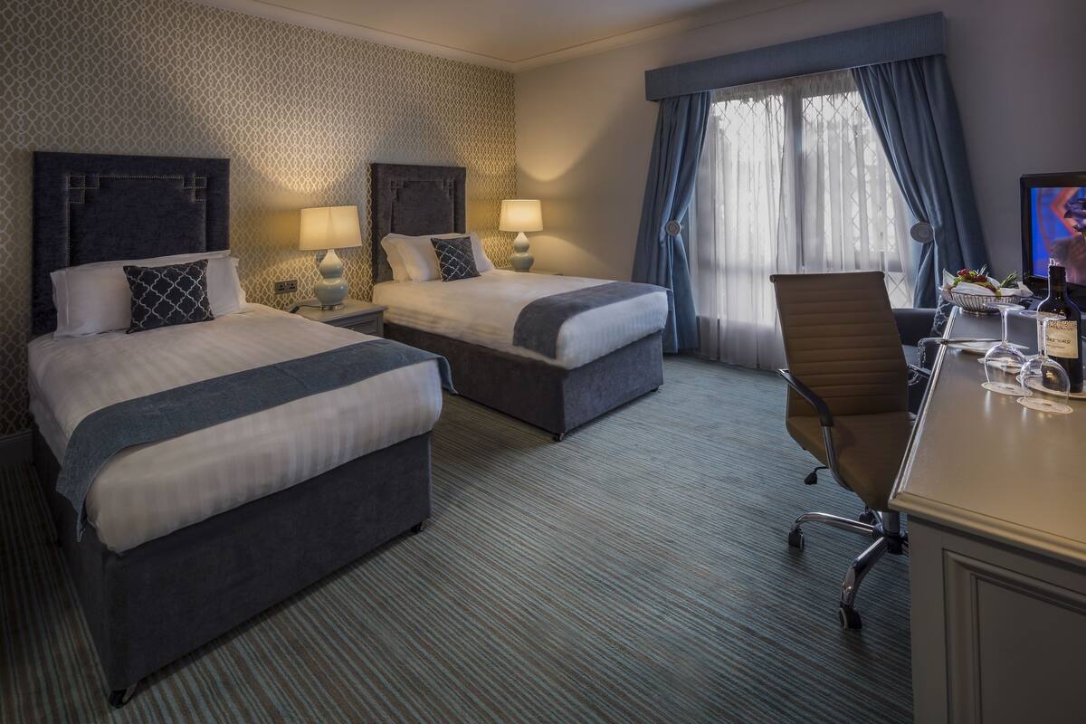 A hotel room with two beds, a desk, a chair, a TV, and a window with curtains.