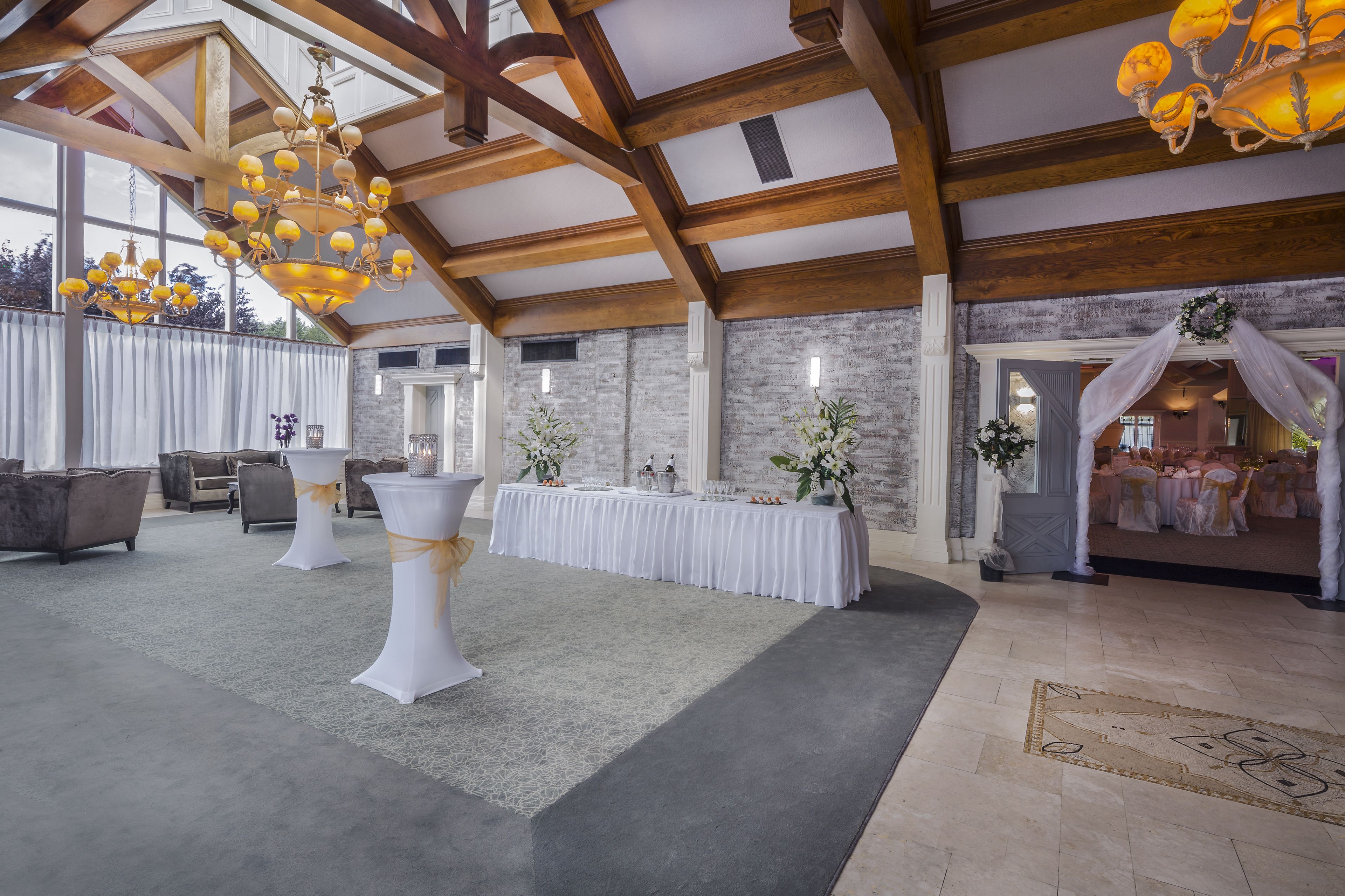 A spacious hall with high ceilings and exposed wooden beams, featuring a long table with white tablecloths and floral centerpieces, and a chandelier with multiple lights.
