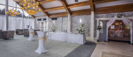 A spacious hall with high ceilings and exposed wooden beams, featuring a long table with white tablecloths and floral centerpieces, and a chandelier with multiple lights.