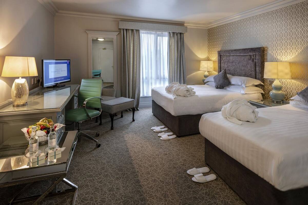 A hotel room with two beds, a desk with a computer, a chair, a small table with toiletries, and a view of the outside through a window.