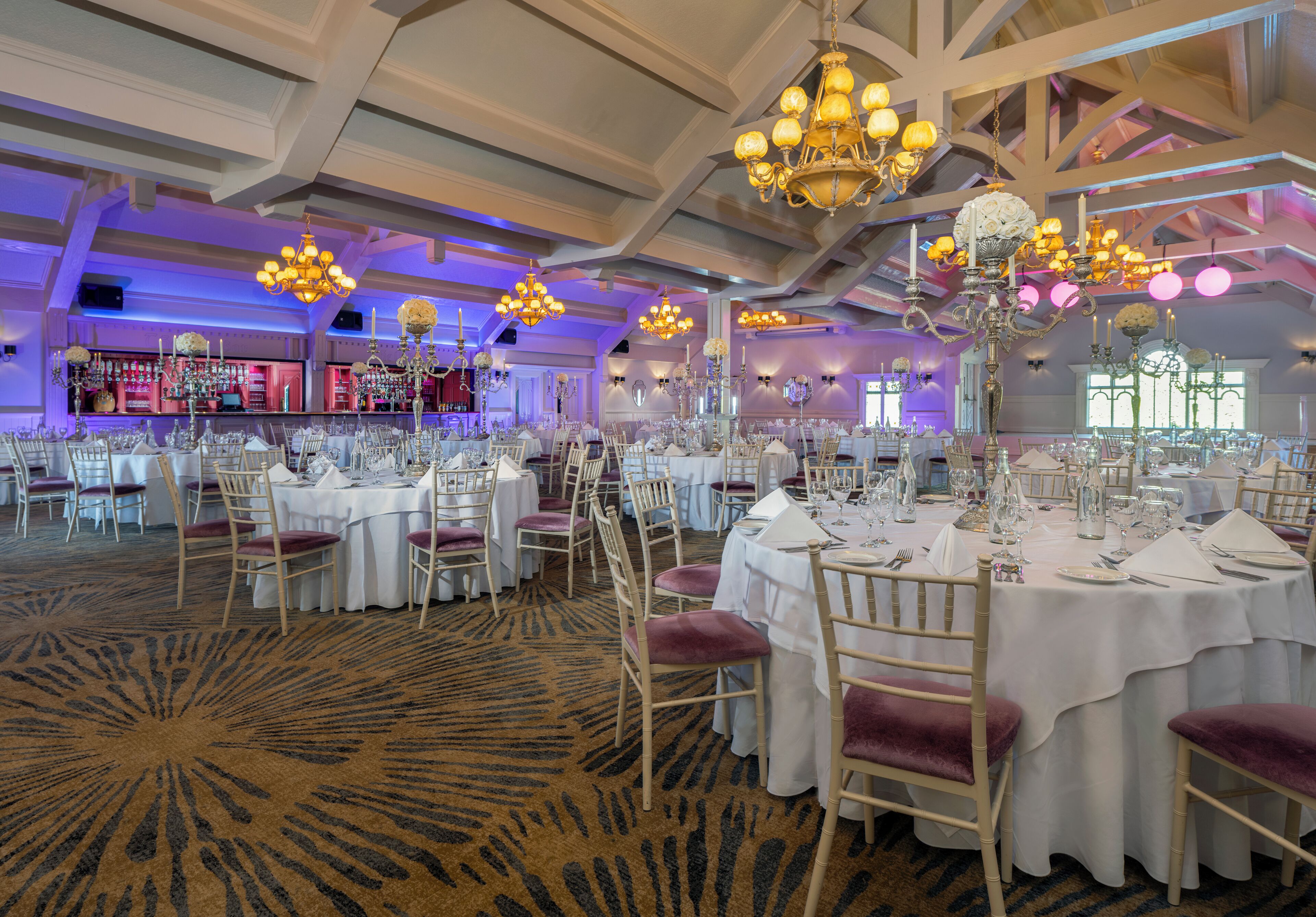 Un salón de banquetes con mesas redondas dispuestas para comer, candelabros y una zona de bar al fondo.