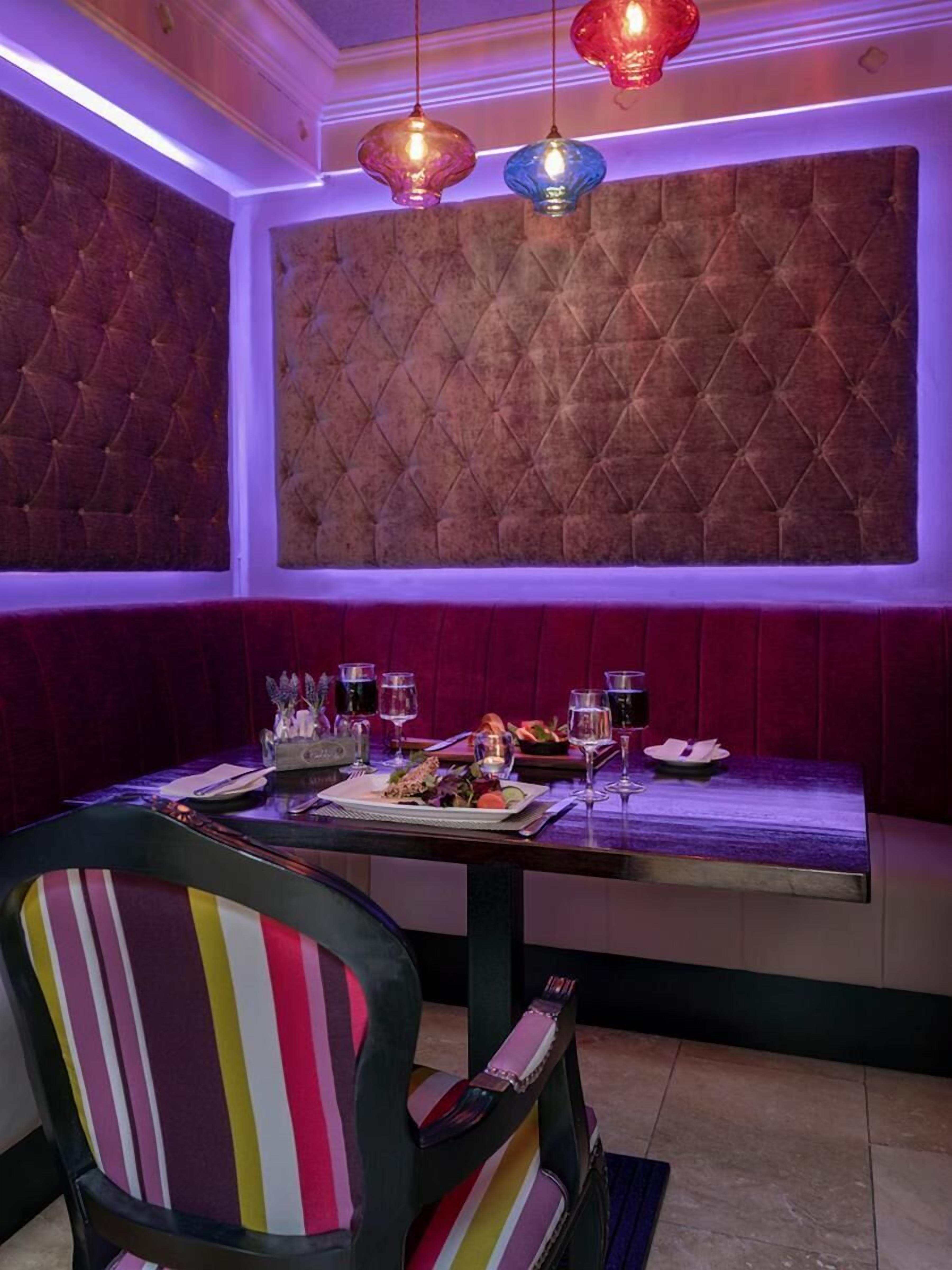 Un comedor con un muro capitoné, una mesa dispuesta con platos, vasos y una vela. Hay sillas coloridas y lámparas colgantes sobre la mesa.