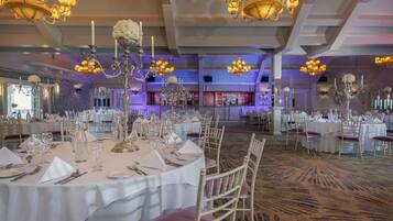 A banquet hall with round tables set for dining, chandeliers, and a bar area in the background.