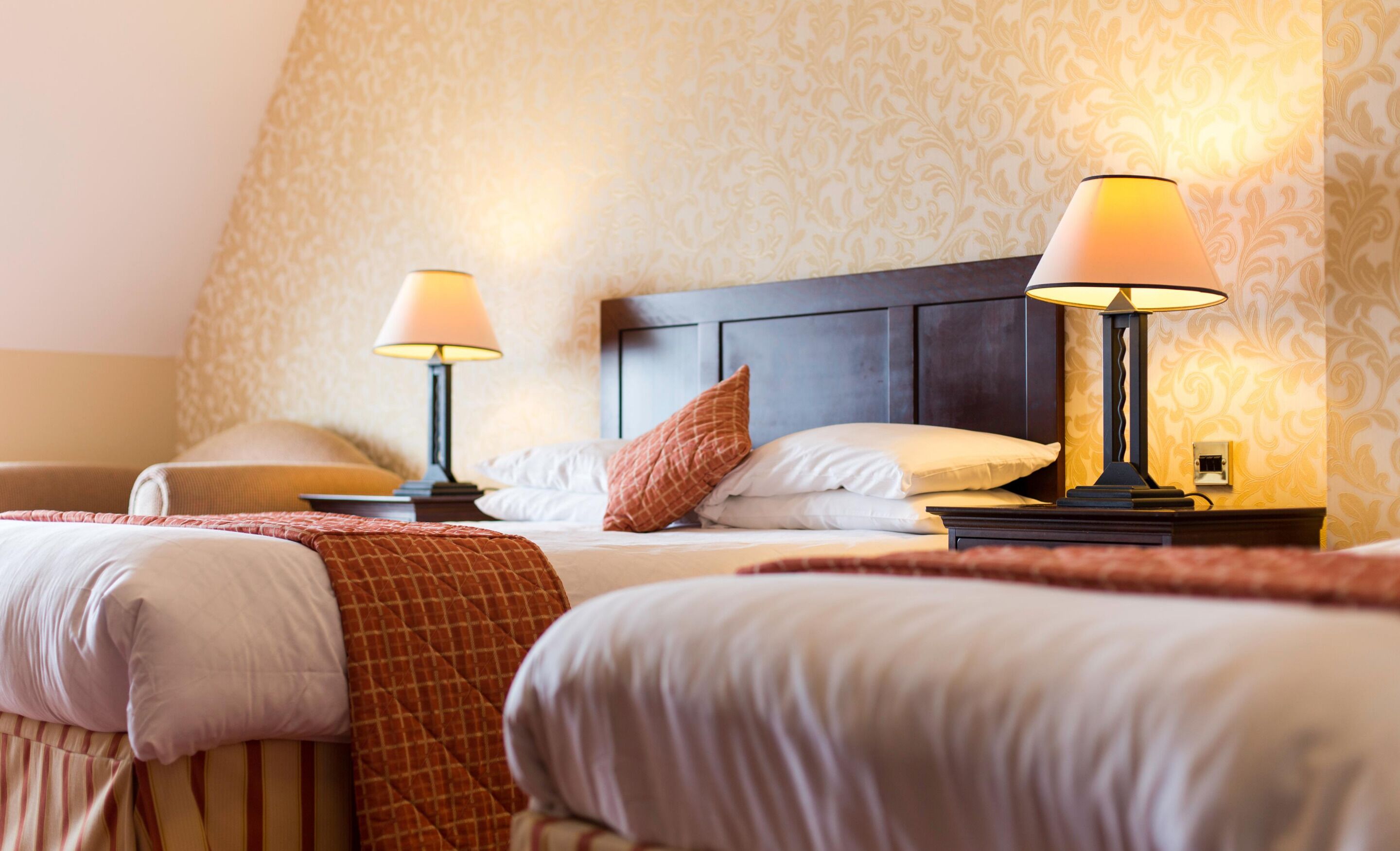 A hotel room with two beds, each with a bedside table and lamp, and a patterned wallpaper.