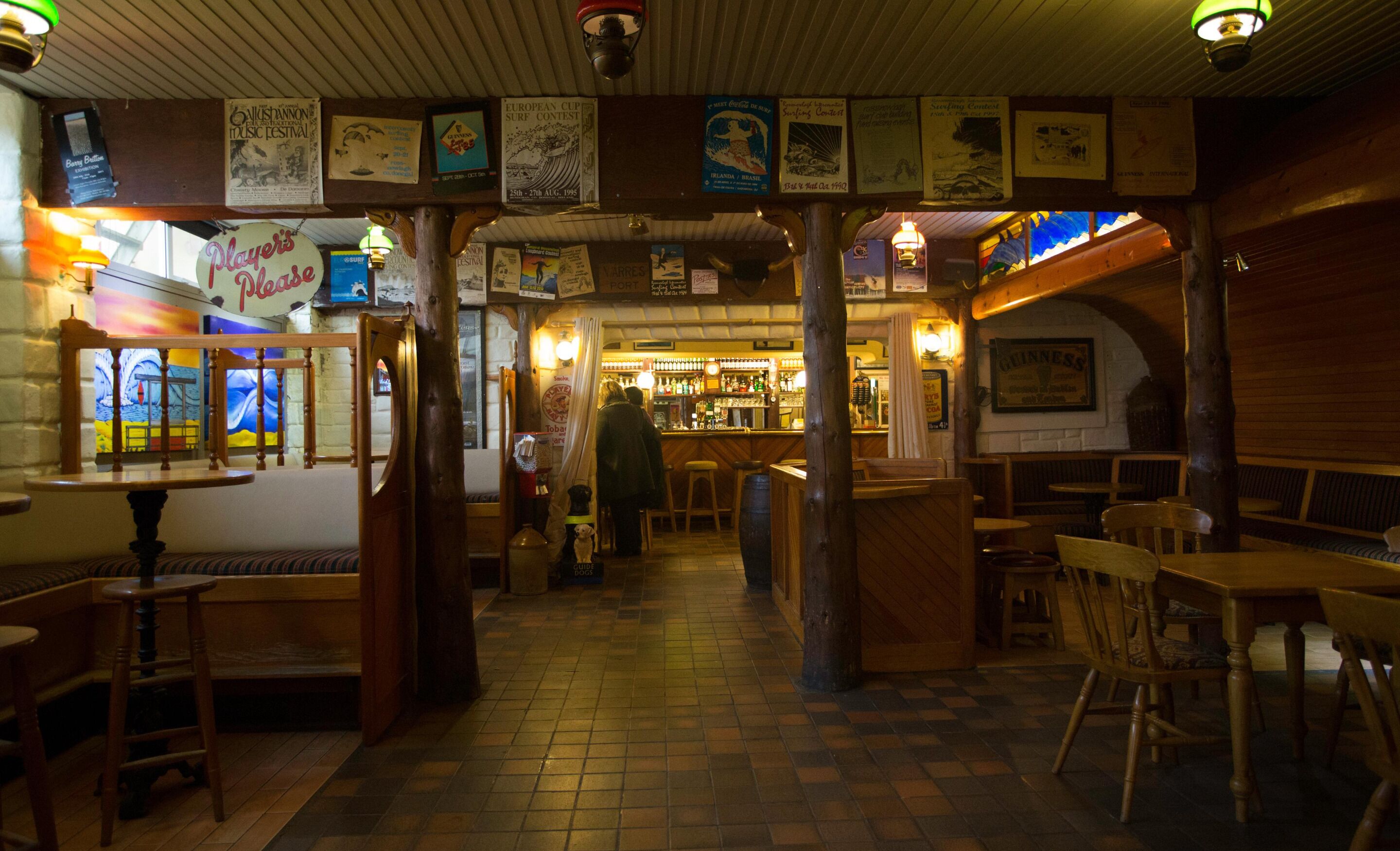 A cozy bar area with wooden furniture, a visible bar counter, and various posters on the walls.