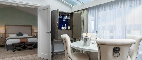 Una habitación de hotel con cama, zona de comedor con mesa y sillas, minibar y un amplio ventanal con cortinas.