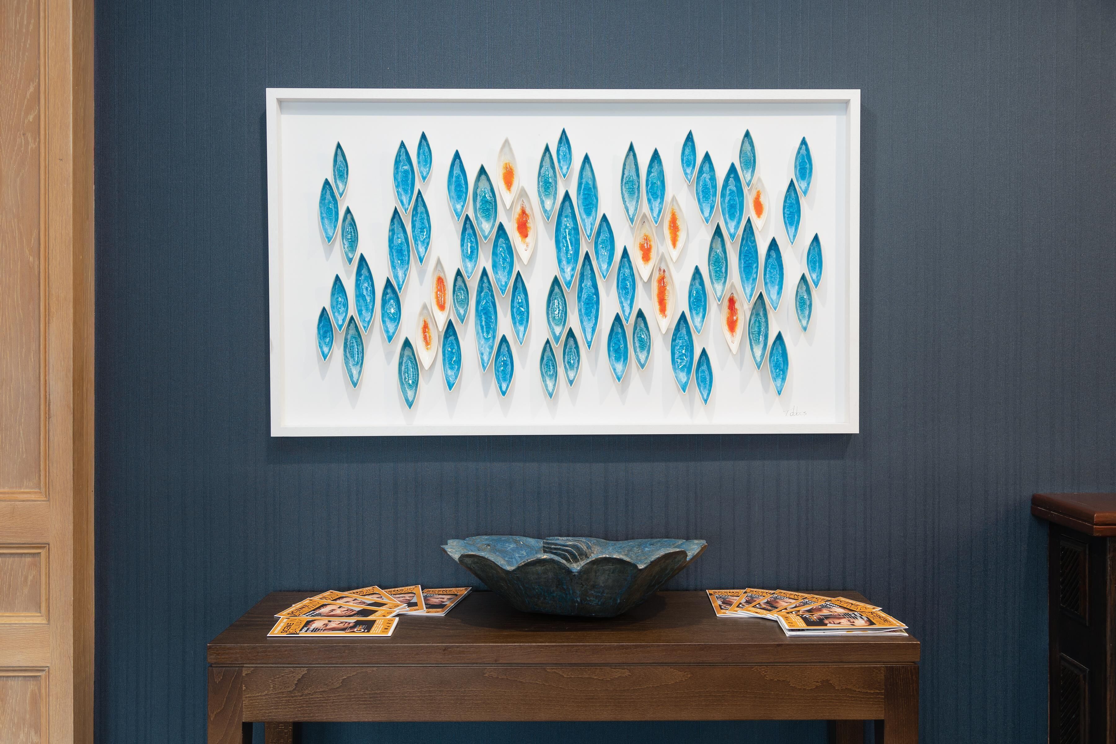 A framed artwork of blue and orange teardrop shapes on a wall, a wooden table with a bowl and magazines, and a dark blue wall.