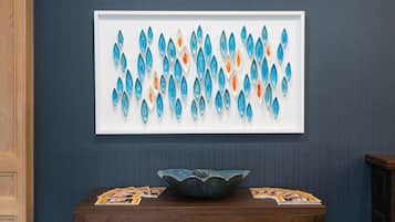 A framed artwork of blue and orange teardrop shapes on a wall, a wooden table with a bowl and magazines, and a dark blue wall.