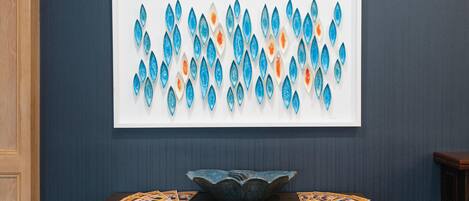 Una obra de arte enmarcada con formas de lágrima azules y anaranjadas en una pared, una mesa de madera con un cuenco y revistas, y una pared azul oscuro.