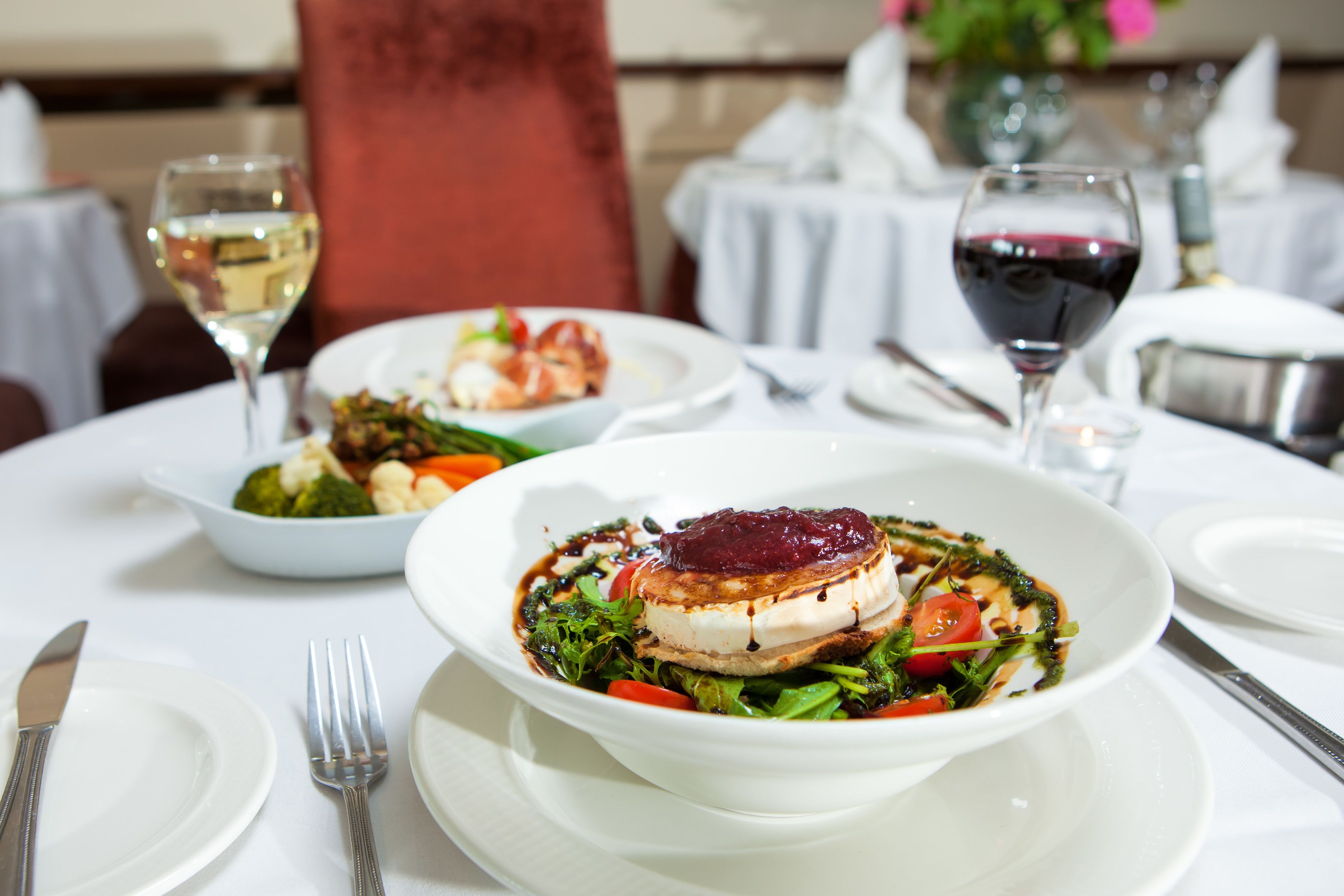 A dining table with white tablecloths, plates, cutlery, and two glasses of wine.