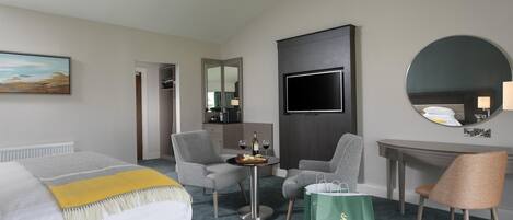 Quarto de hotel com cama, duas poltronas, uma mesinha de centro com garrafa e copo, escrivaninha com cadeira, televisão e um espelho grande.