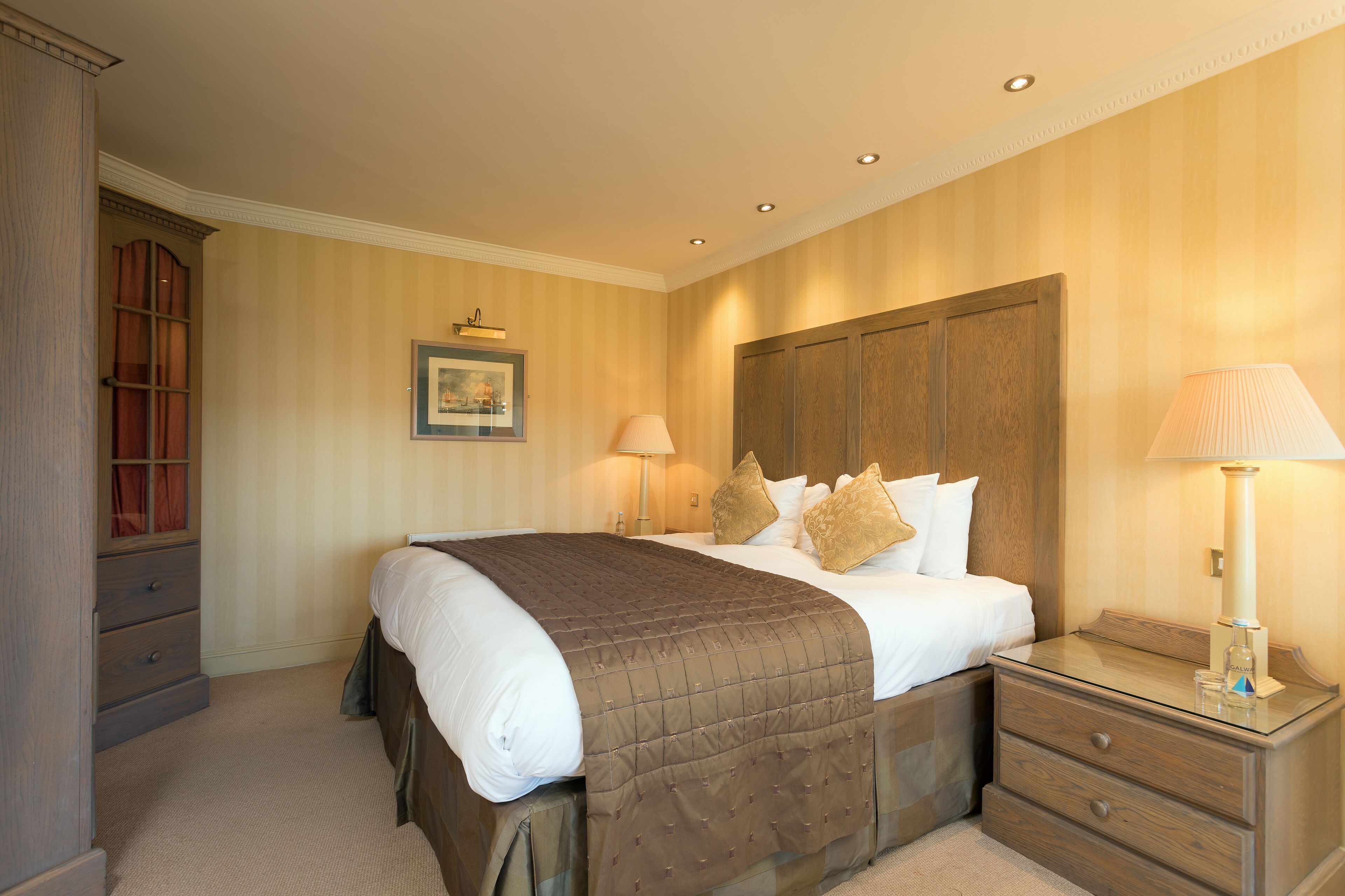 A neatly arranged bedroom with a large bed, a bedside table with a lamp, a wooden wardrobe, and a framed picture on the wall.