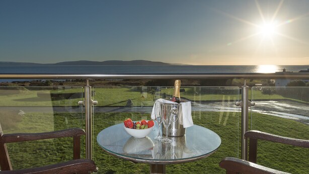 A glass table with a bottle of champagne, a bowl of fruit, and two glasses on a balcony overlooking a body of water and distant hills. - Connemara Coast Hotel (Galway)