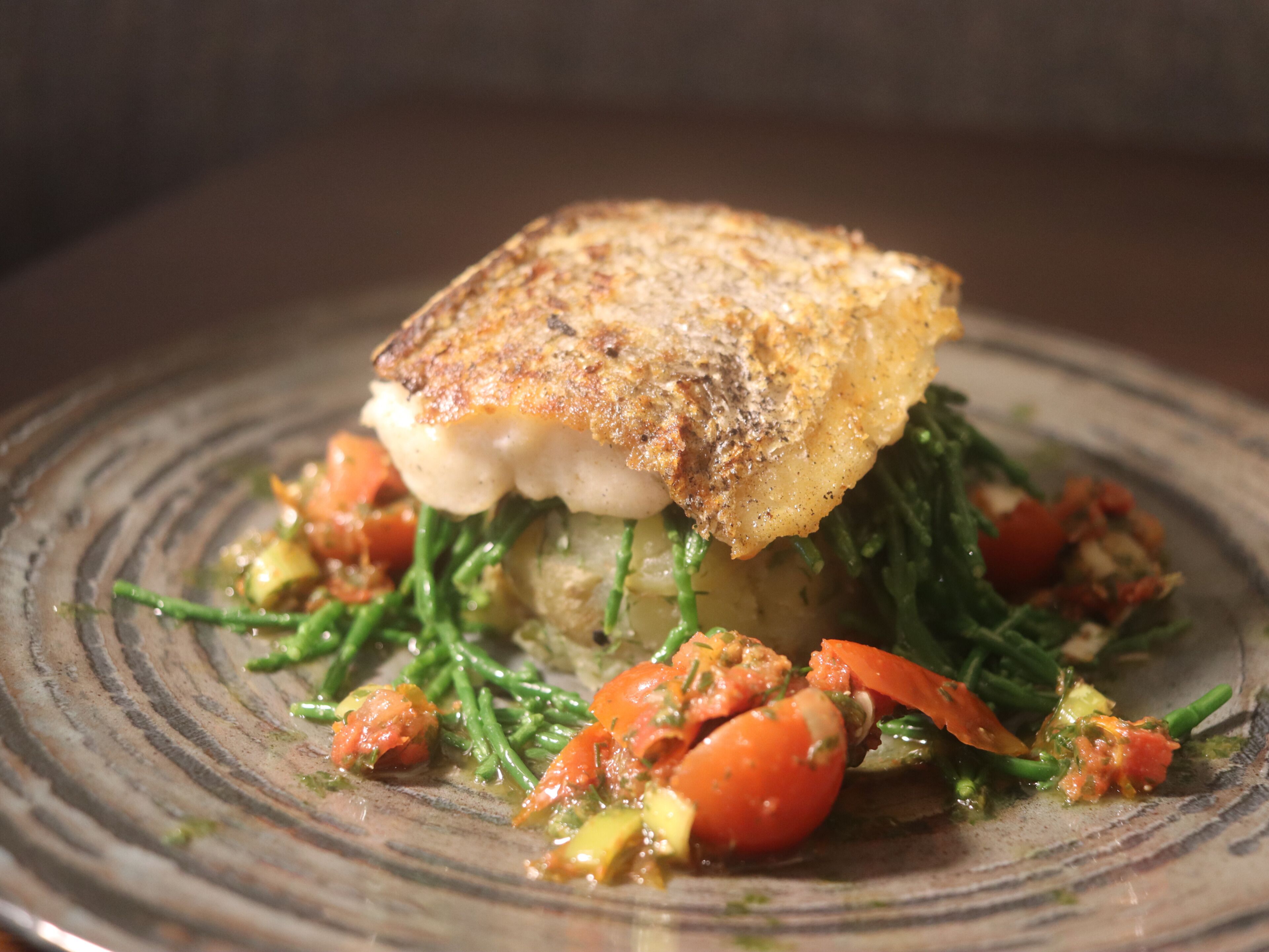 A plate with a piece of fish topped with a golden-brown crust, accompanied by green beans, tomatoes, and a sauce.