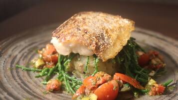 Un plato con un trozo de pescado cubierto de una costra dorada, acompañado de judías verdes, tomates y salsa.