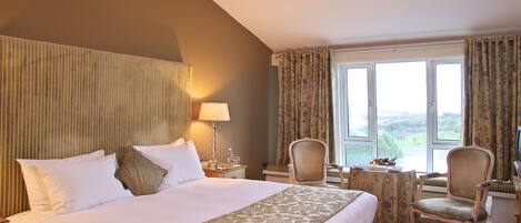 A hotel room with a large bed, a desk with a chair, a lamp, and a view of the outdoors through a window.