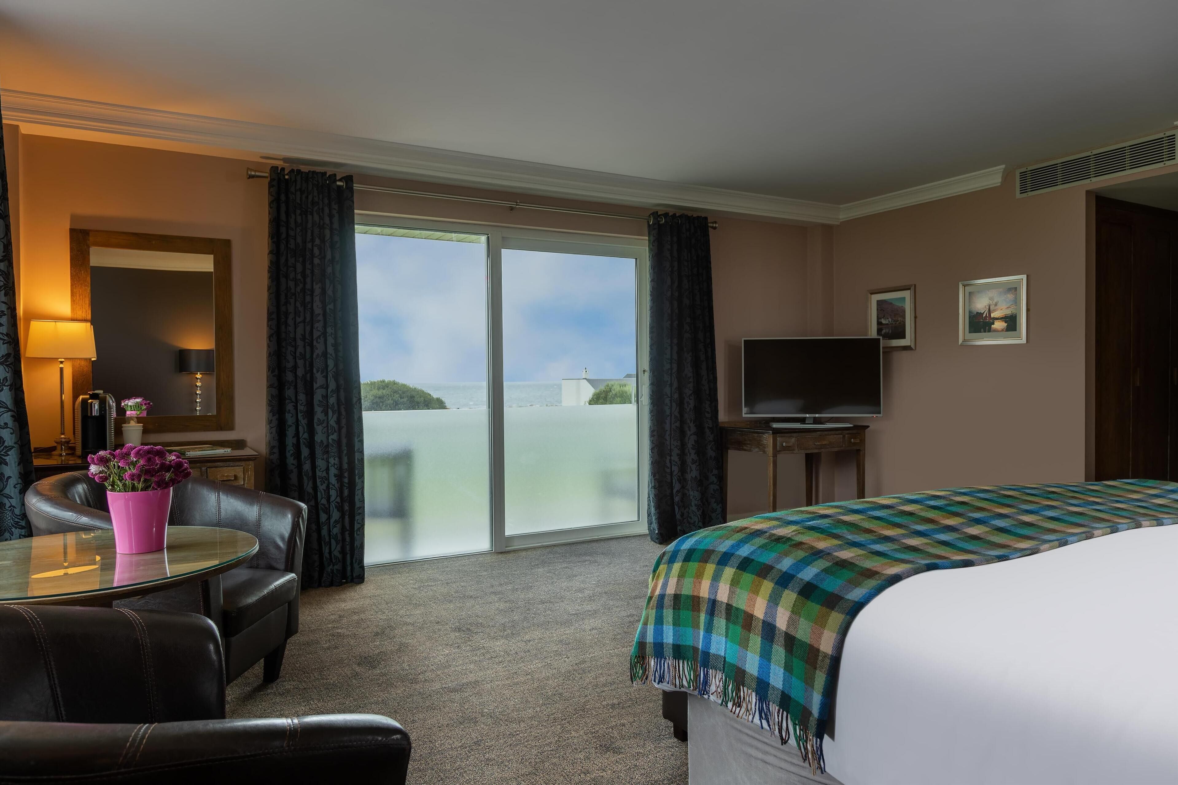 Habitación de hotel con cama, televisor, escritorio con lámpara, silla, mesa de vidrio con un florero y vista al mar a través de puertas correderas amplias.