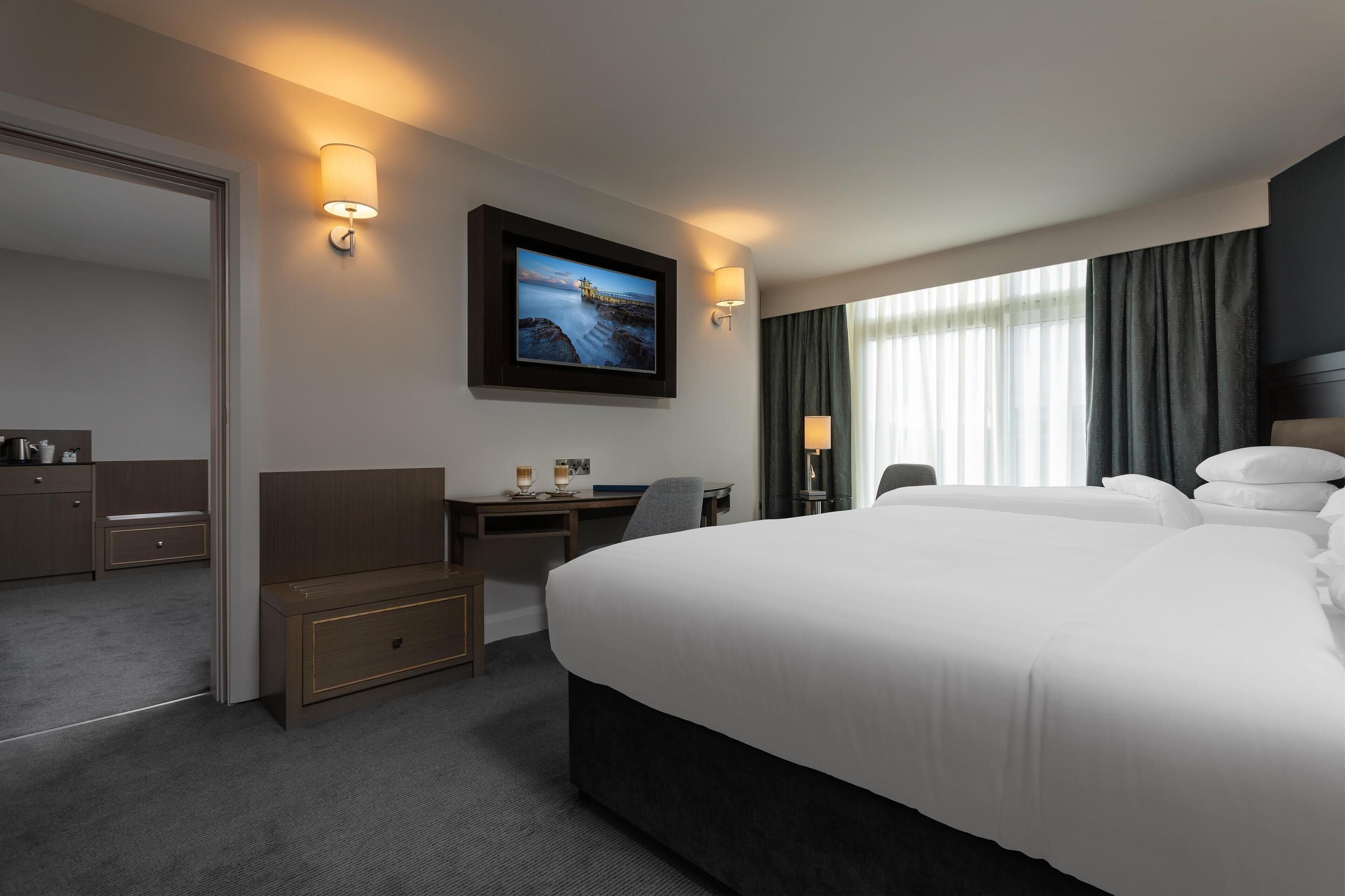 A hotel room with a large bed, a desk with a chair, a television mounted on the wall, and a window with curtains.