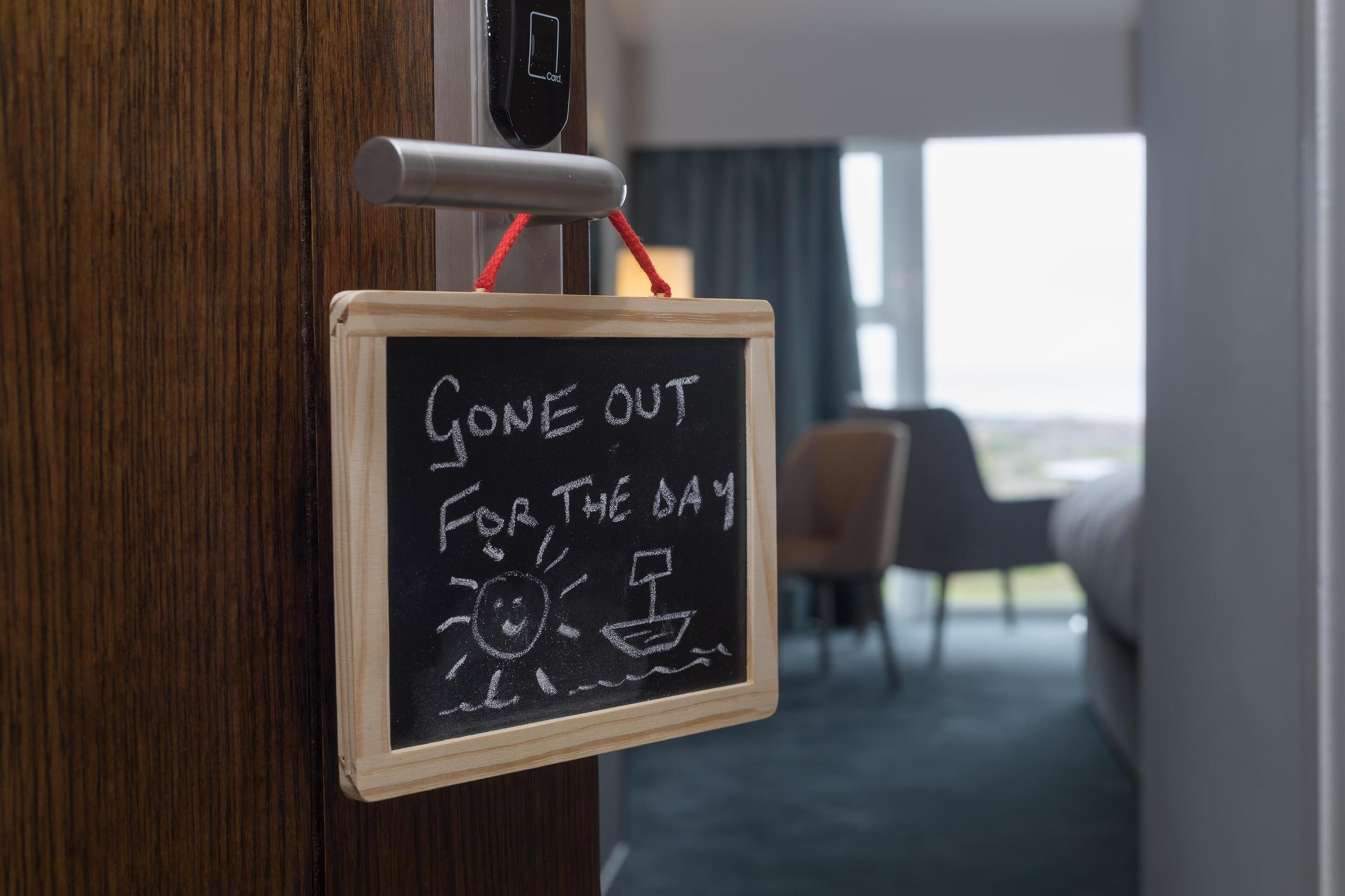 A hotel room with a chalkboard sign on the door that reads "Gone out for the day" and a small drawing of a sun and a boat.