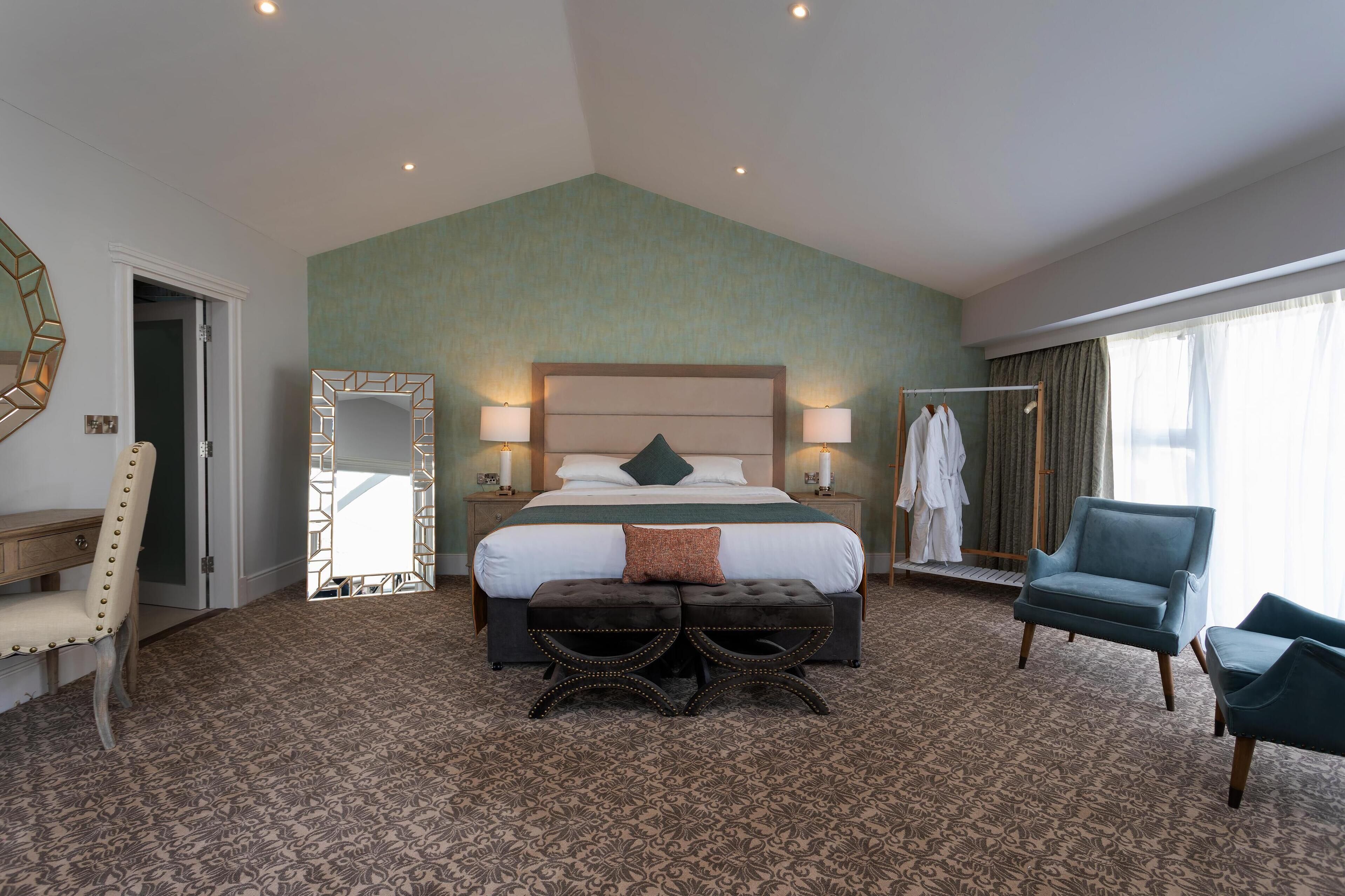 A hotel room with a large bed, a sitting area with a chair and ottoman, a mirror, a wardrobe, and a window with curtains.