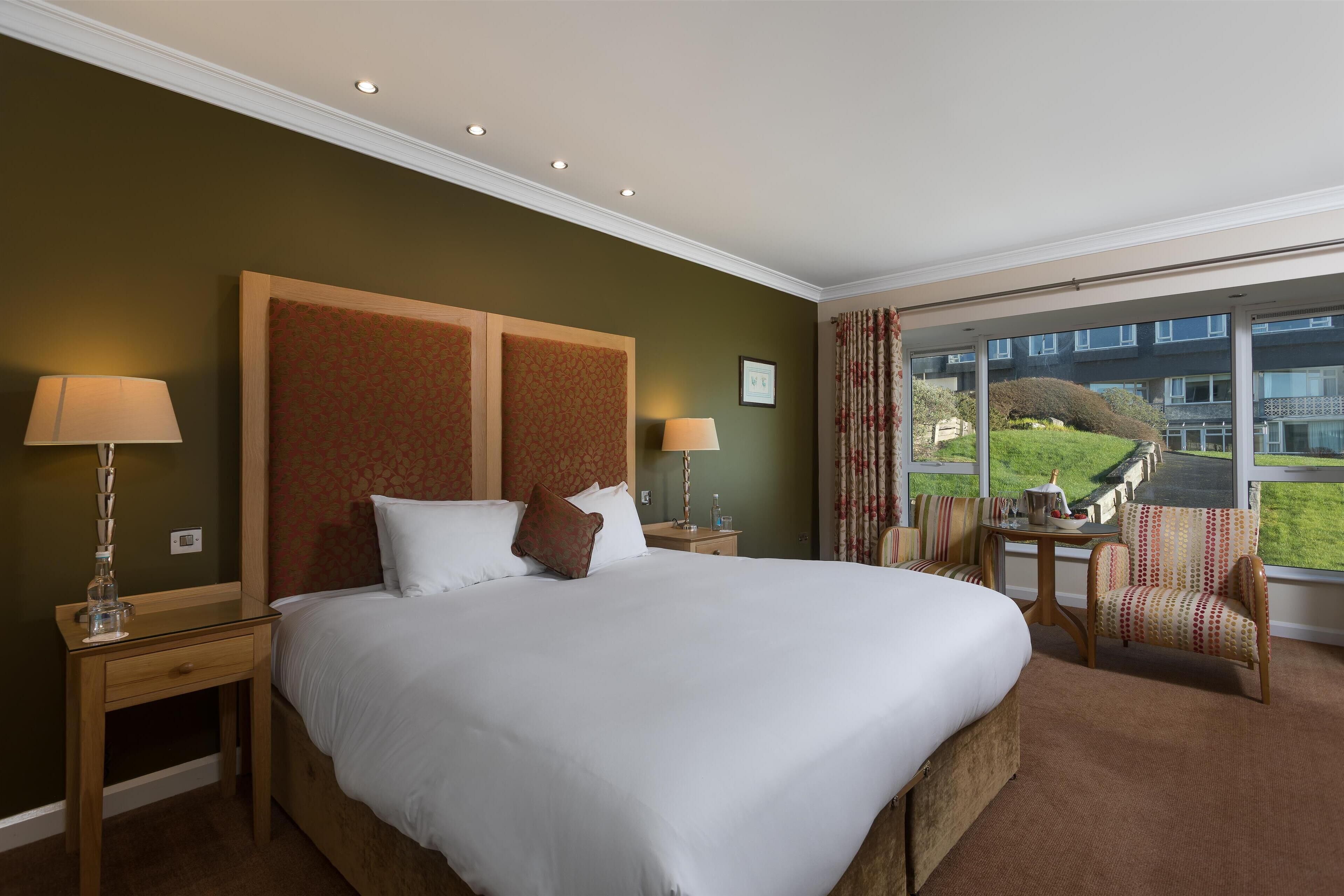 Ein Hotelzimmer mit einem großen Bett, zwei Nachttischen, einem Sessel und Blick auf ein Gebäude sowie Grünflächen.