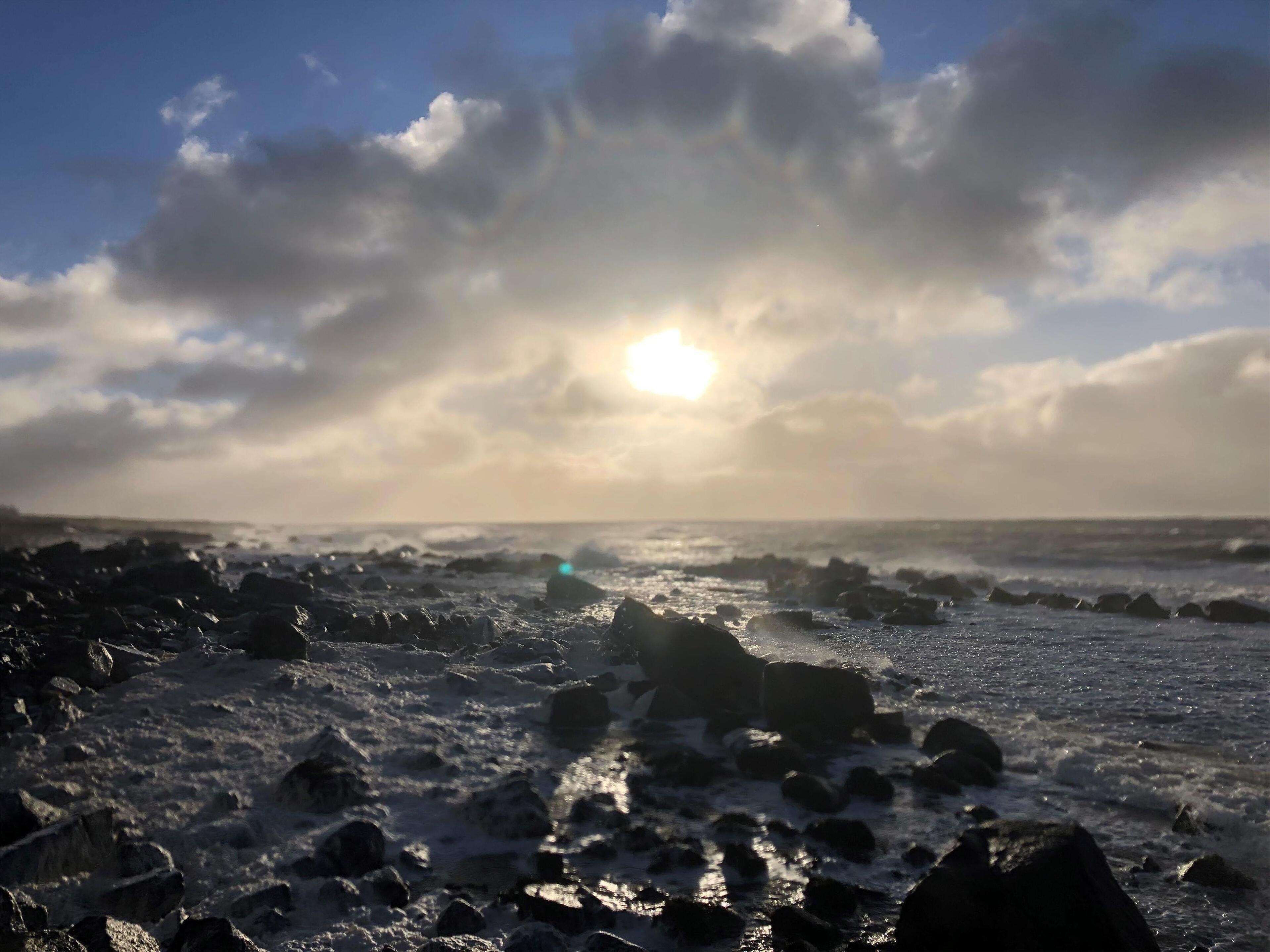 A rocky shoreline with waves crashing against the rocks under a partly cloudy sky with the sun visible.