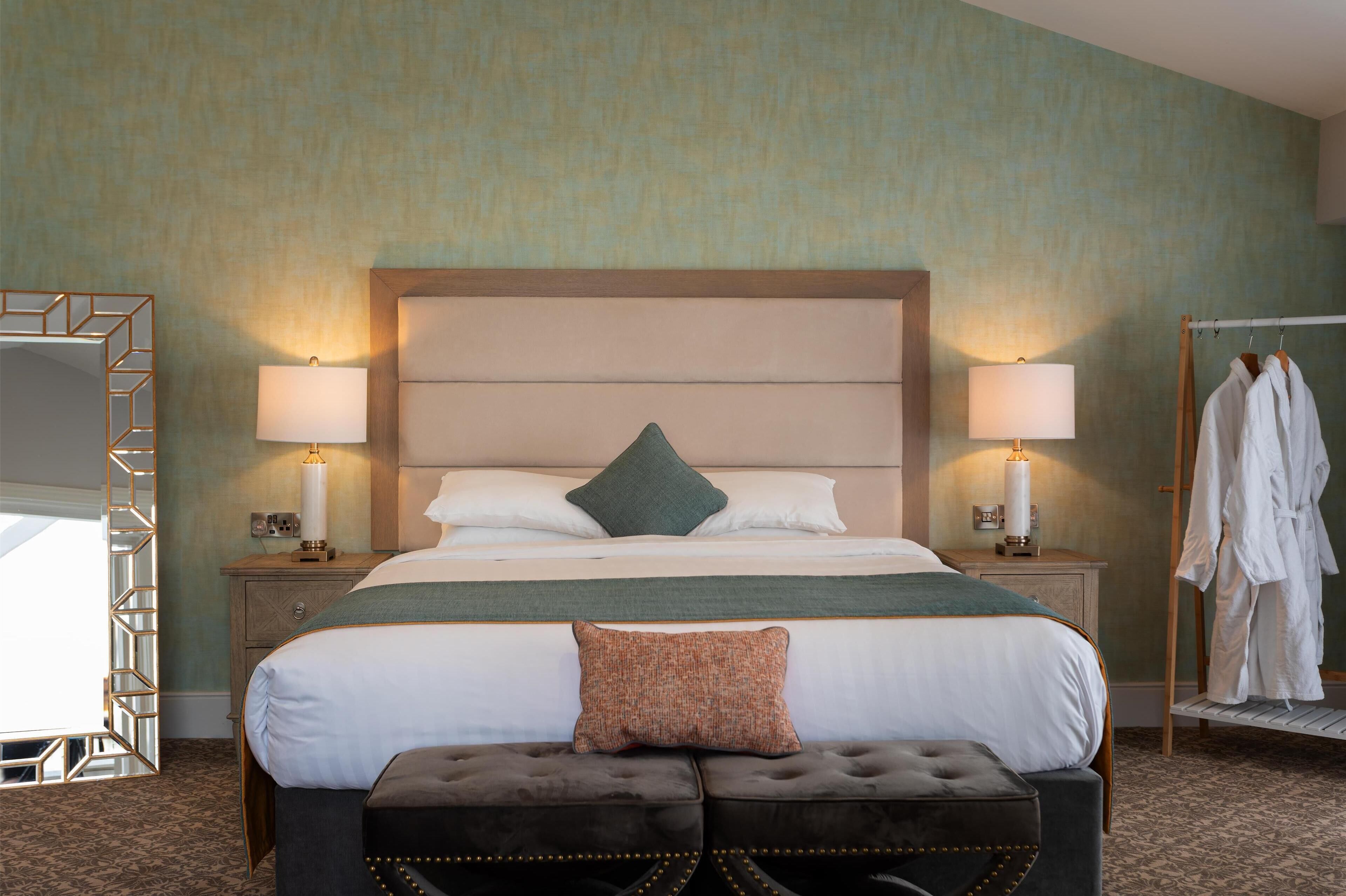 A hotel room with a large bed, two bedside lamps, a mirror, a robe rack, and an ottoman.