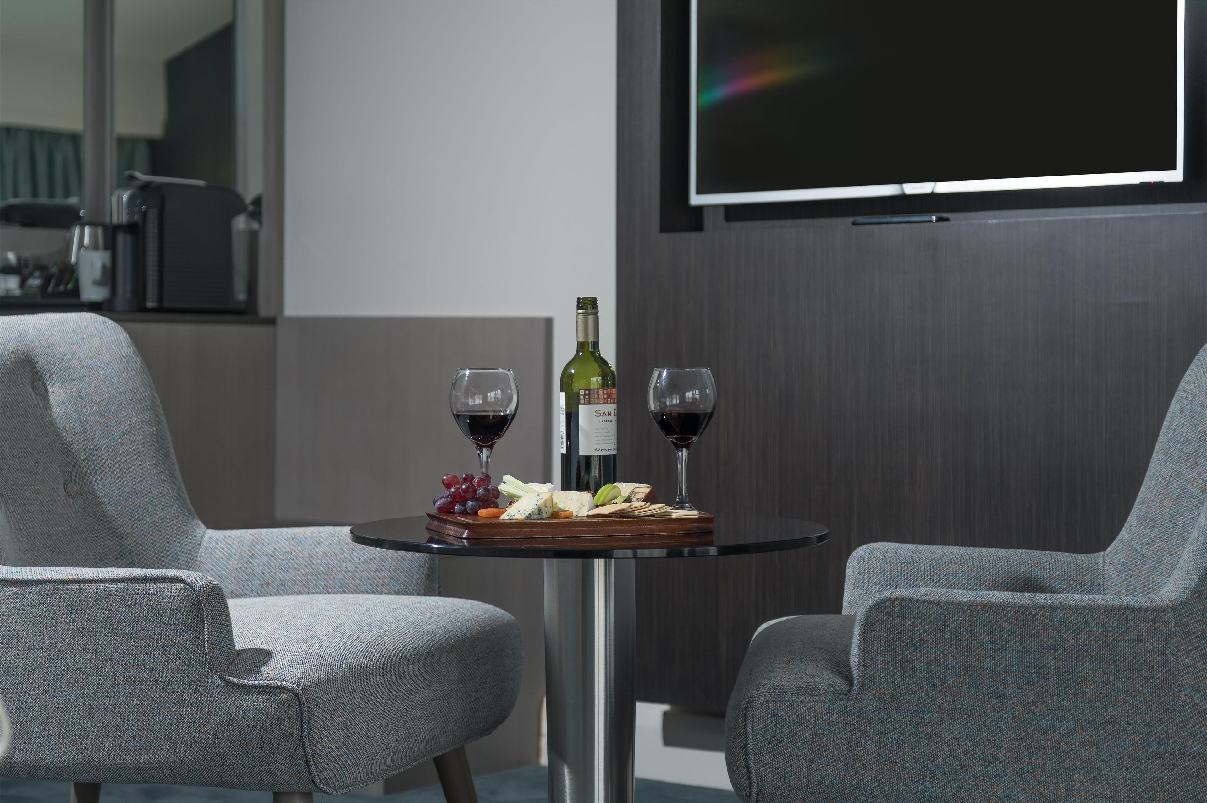 Une pièce moderne avec deux fauteuils gris à revêtement en tissu, une petite table ronde sur laquelle se trouvent une bouteille de vin, des verres et une planche à fromages.