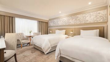 Quarto de hotel com duas camas, uma escrivaninha, uma cadeira e uma mesinha.